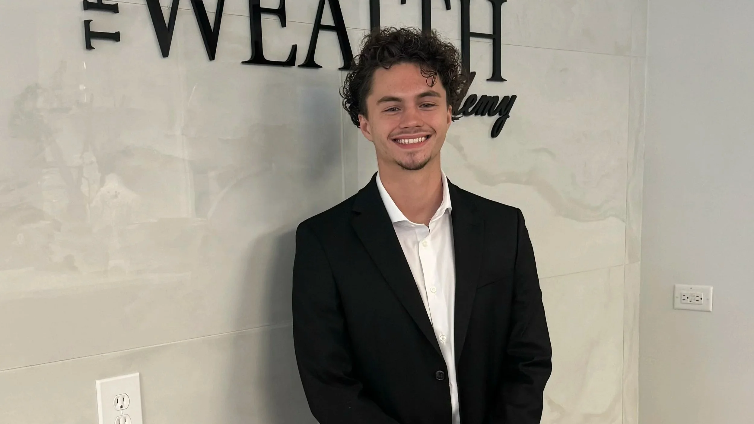 A young man with curly dark hair, wearing a black suit and white shirt, standing in front of a white wall with black letters that spell out a word, and smiling at the camera.
