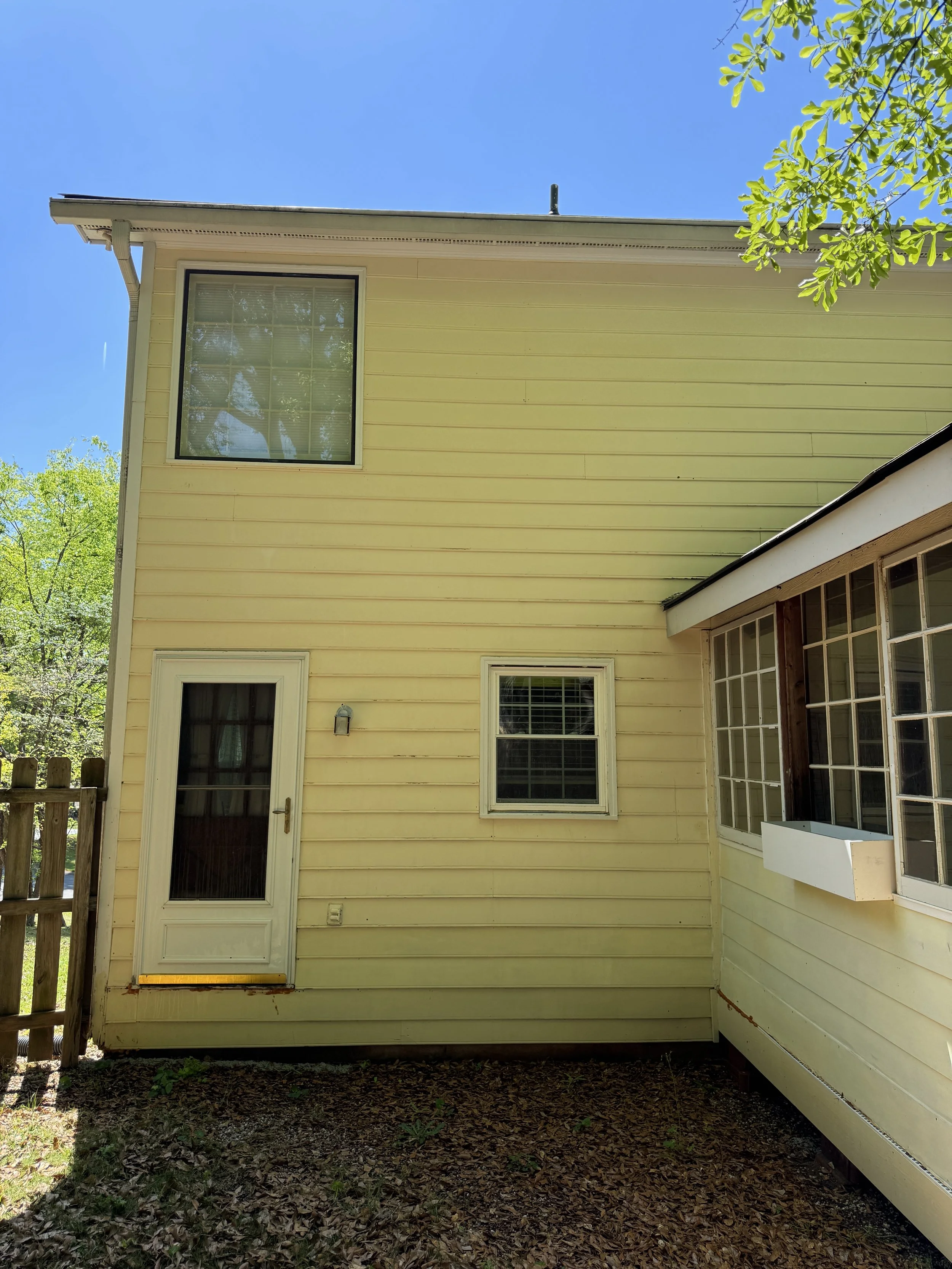 A yellow two-story house with three windows, a door, and an overhang on the right side, surrounded by greenery and a blue sky.