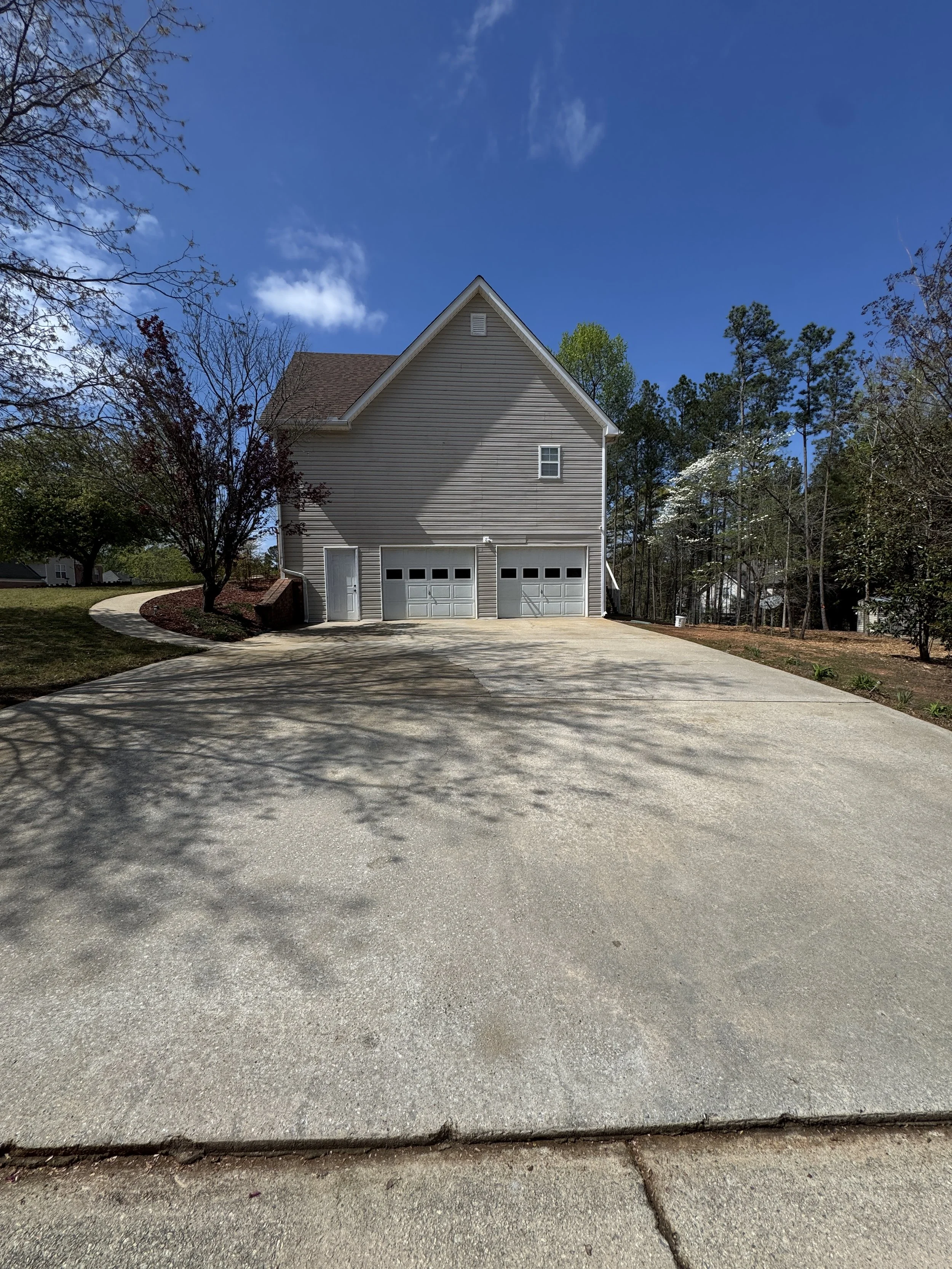 A residential driveway leading to a house with a two-car garage, surrounded by trees and a blue sky with some clouds.