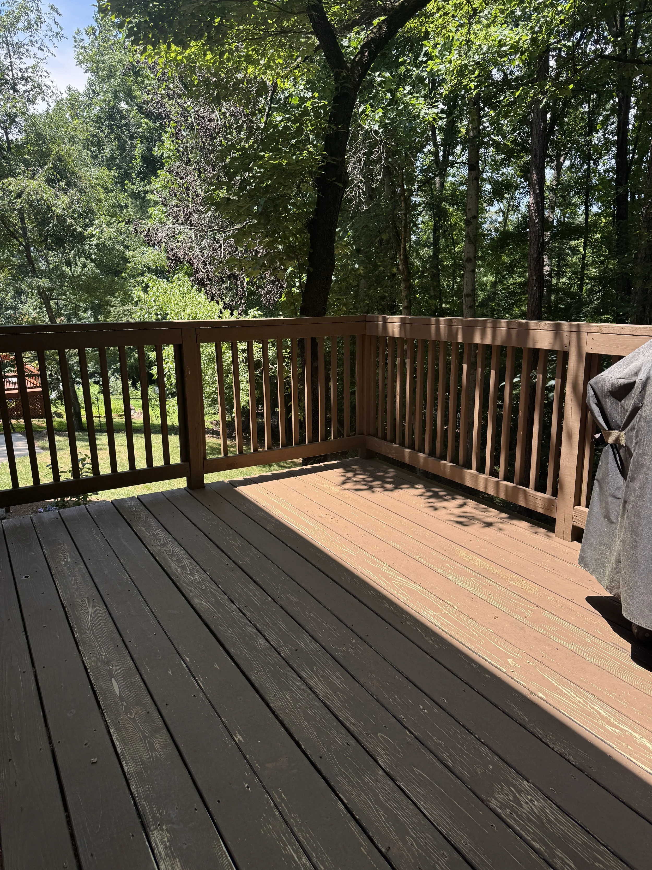 Wooden deck with railing overlooking a wooded backyard with green trees and grass, partially shaded by tree branches.
