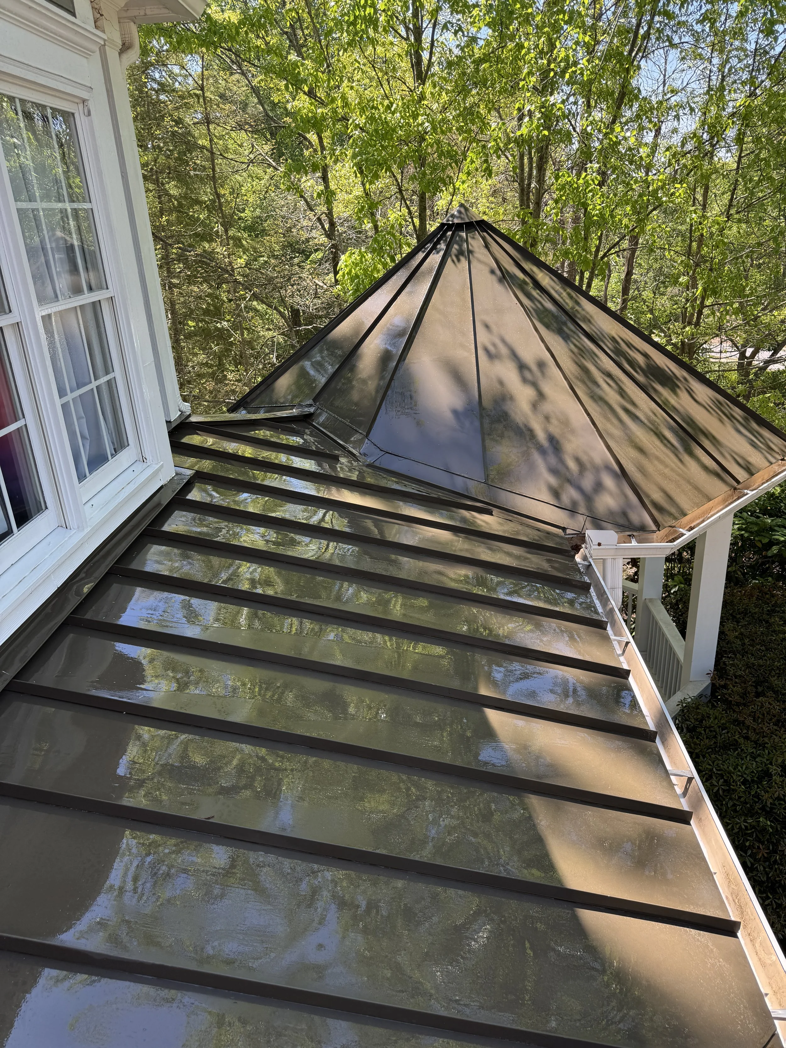 viewed from a house window, a metal roof with a pyramid-shaped cap, surrounded by trees with green leaves.