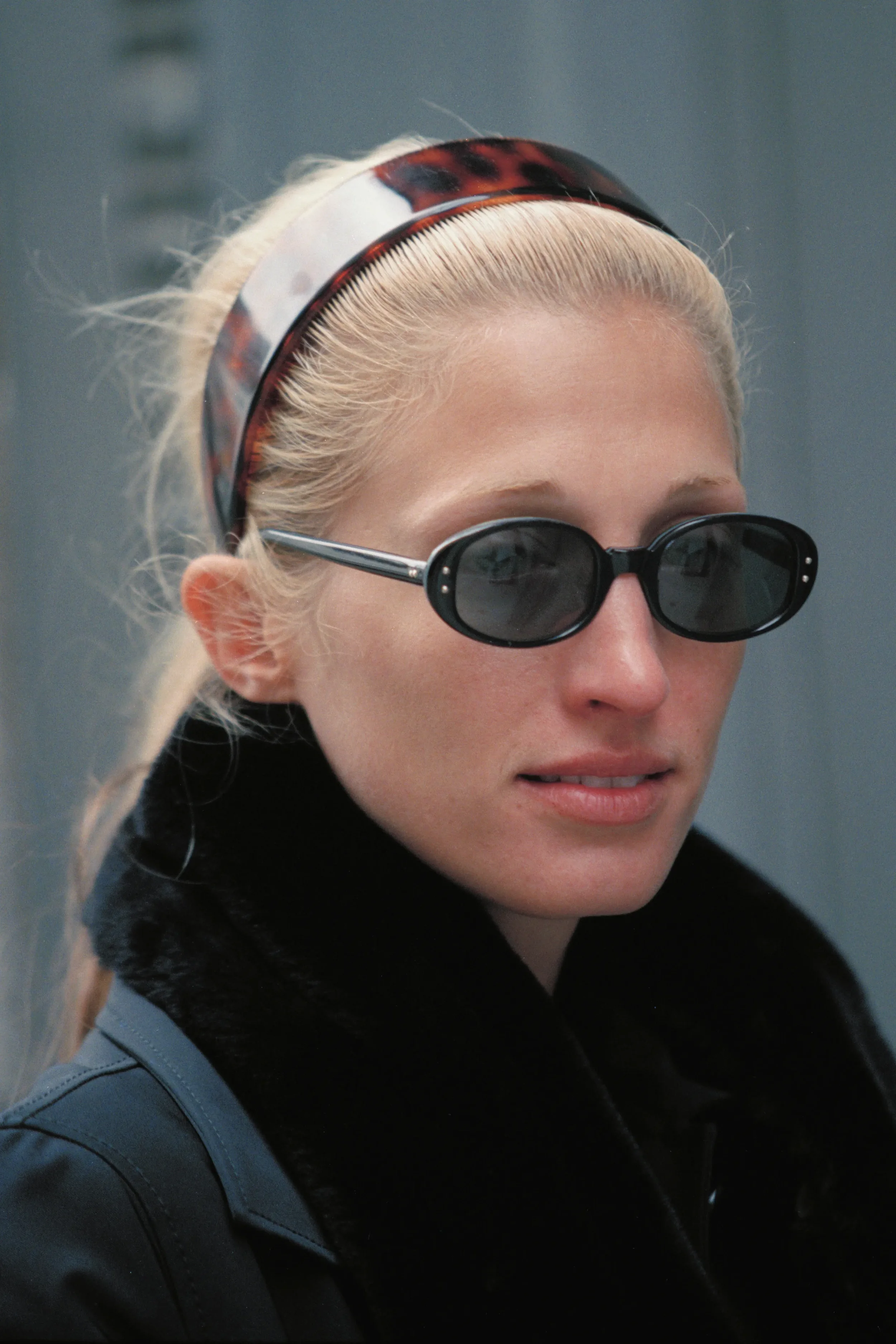 Close-up of a woman with blonde hair, wearing black sunglasses, a tortoiseshell headband, and a black coat with a fur collar.