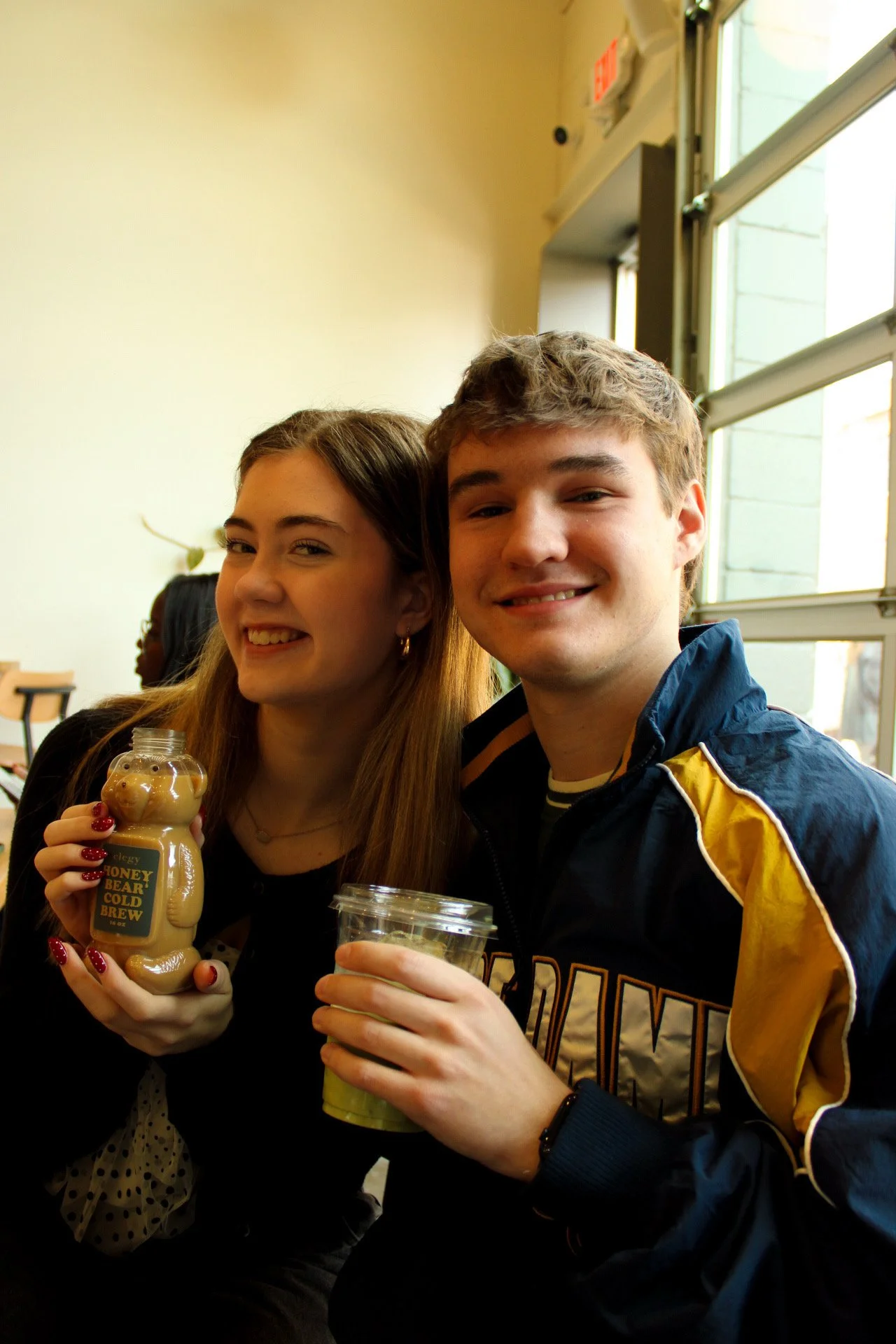 Two smiling teenagers, a girl and a boy, sitting in a sunlit room with large windows. The girl is holding a bottled beverage labeled "Honey Bear Cold Brew."