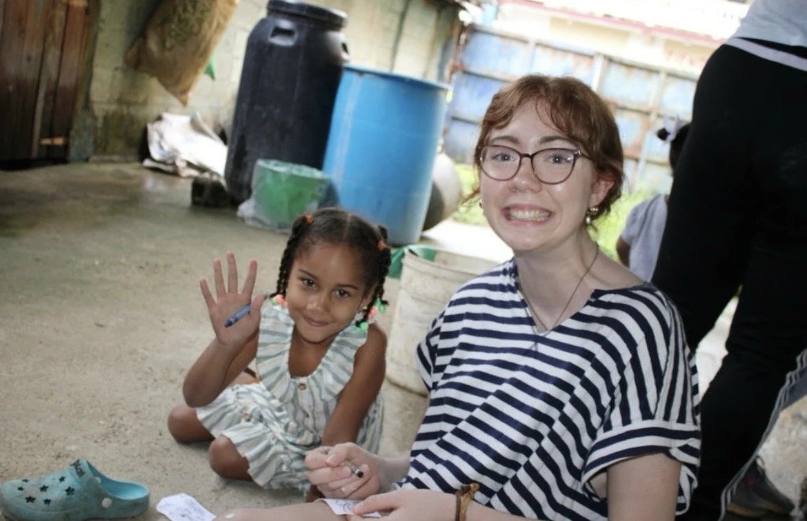A young girl sitting on the ground and a young woman with glasses smiling at the camera. The girl is waving and holding a pencil, wearing a striped dress. The woman is wearing a striped shirt and has short, light brown hair. They are outdoors, with barrels and a fence in the background.