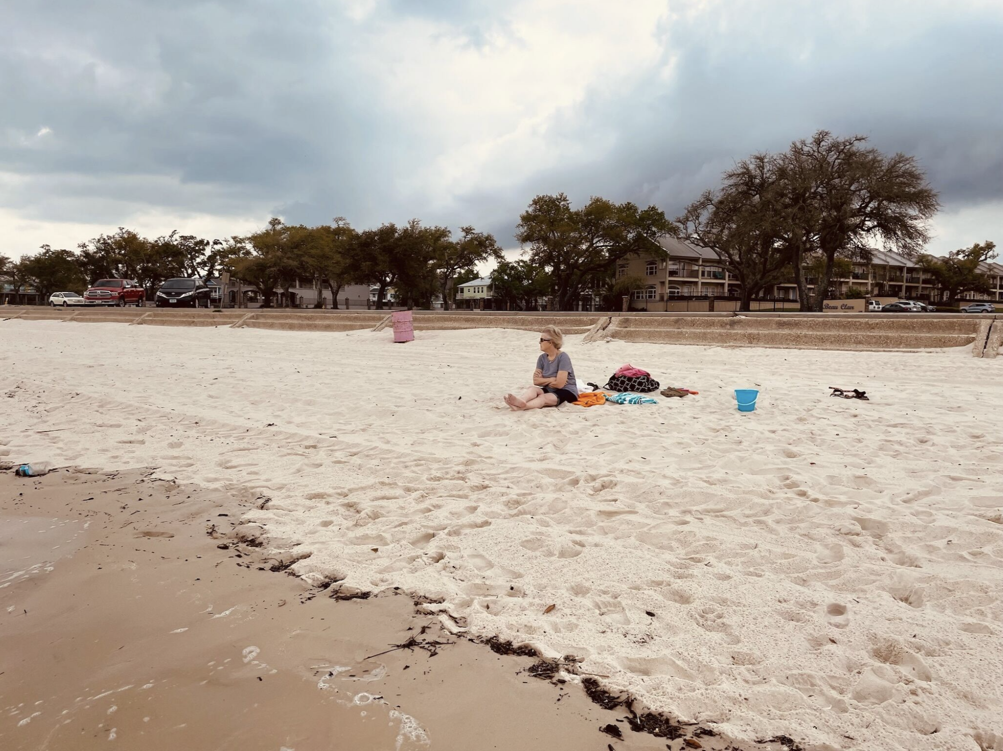 A woman sitting alone on a sandy beach near the water with beach towels, a pink bag, a blue bucket, and small toys nearby. Overcast sky with dark clouds and houses in the background.