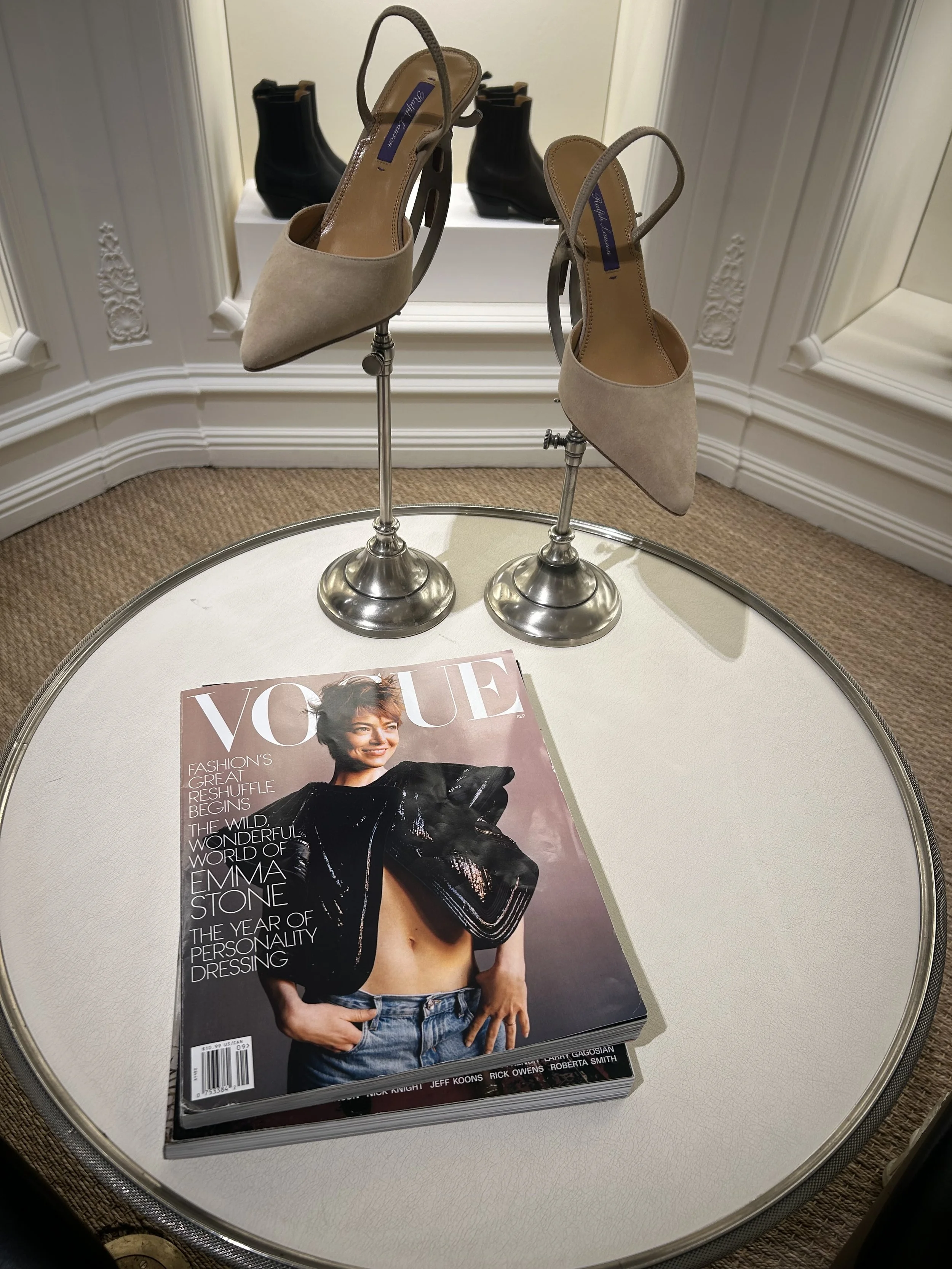 A Vogue magazine on a white table in front of two beige and black high-heeled shoes displayed on metal stands.