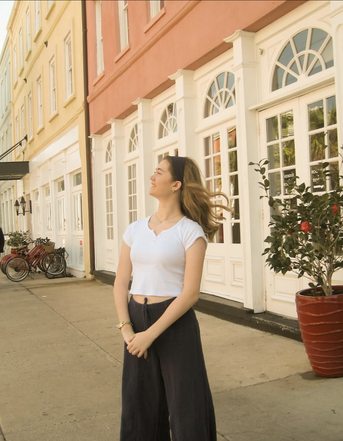 A smiling woman with wavy hair, wearing a black headband, white crop top, and dark wide-leg pants, standing on a sidewalk in front of colorful European-style buildings with large arched windows and potted plants.