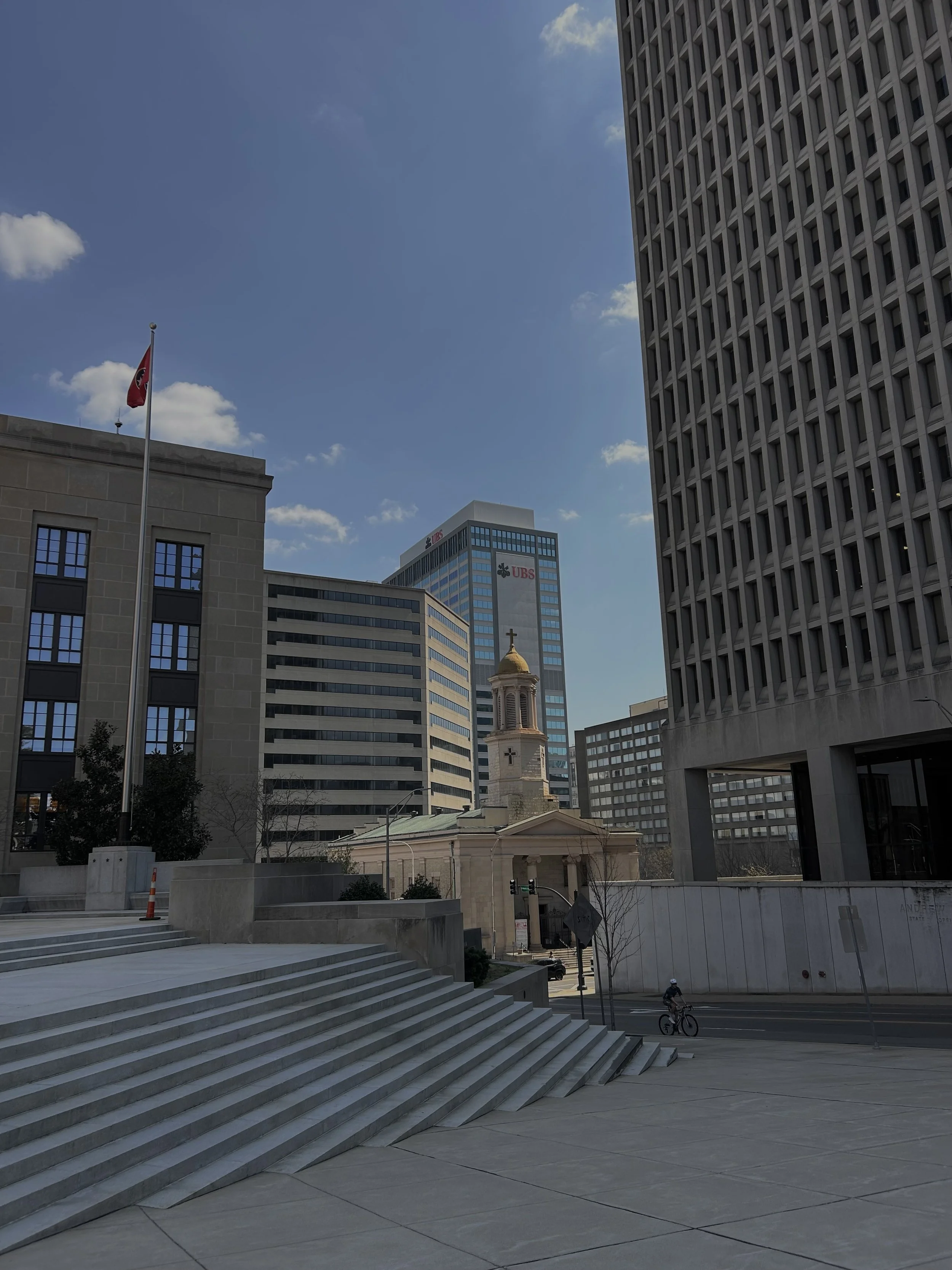 Cityscape with modern buildings, a small church with a cross on top, and a man riding a bicycle on the street.