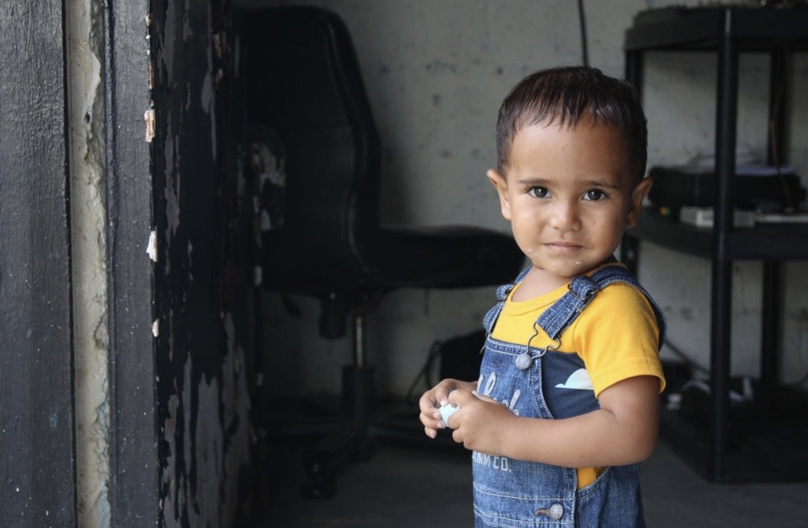 A young boy with short dark hair and brown eyes, wearing a yellow shirt and denim overalls, standing indoors with a serious expression, holding a small object in his hands, in a room with a black office chair and a black shelf in the background.