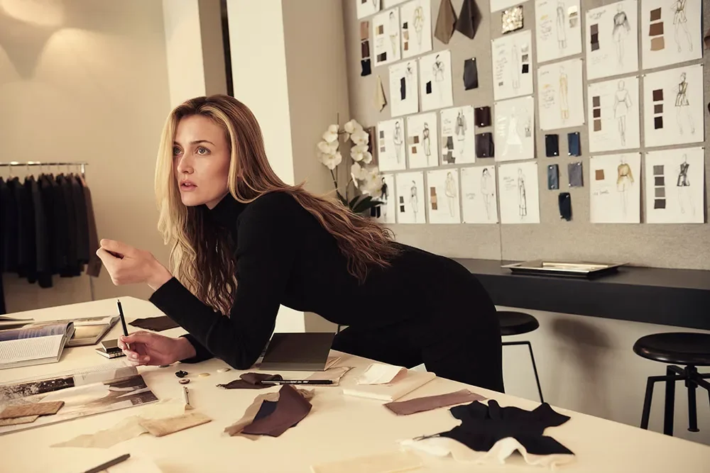 A woman sketching fashion designs at a studio workspace with fabric swatches, pencils, and books, surrounded by design sketches and color palettes on the wall.