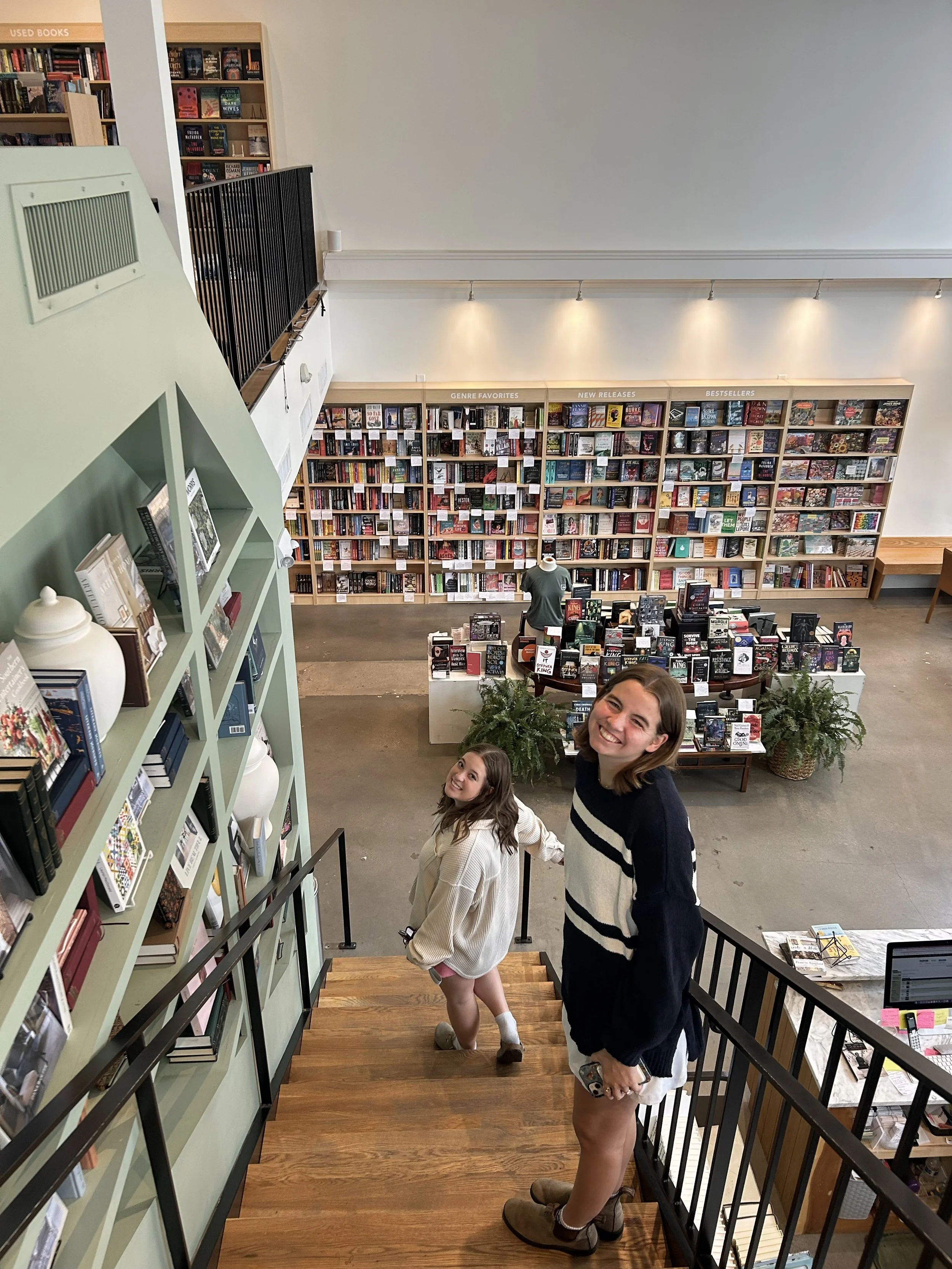 Two young women smiling and looking up while ascending a staircase inside a bookstore with shelves of books on the walls and a display table in the center.
