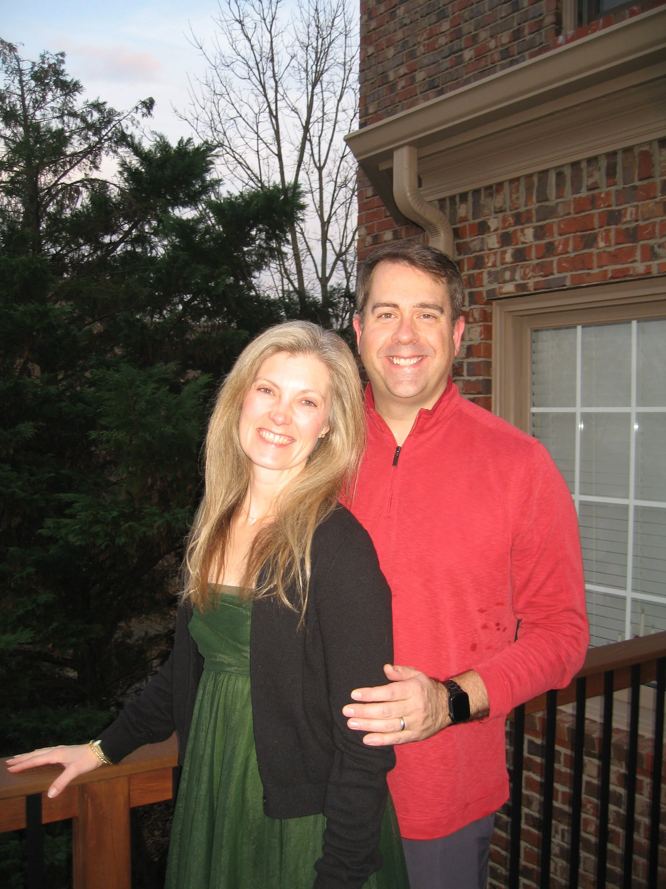 A smiling woman and man standing on a wooden deck outside a brick house with a large window, trees, and a cloudy sky in the background.