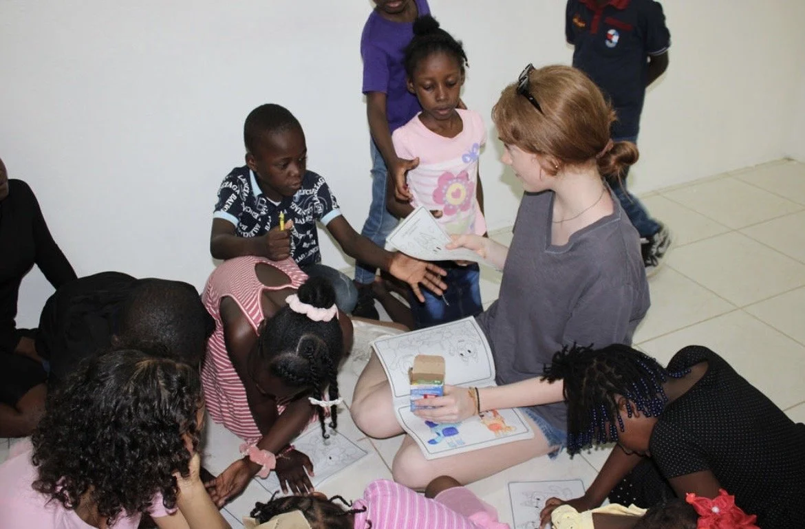 A woman with red hair sitting on the floor surrounded by children, some sitting and some standing, engaging in activities with paper and coloring supplies.