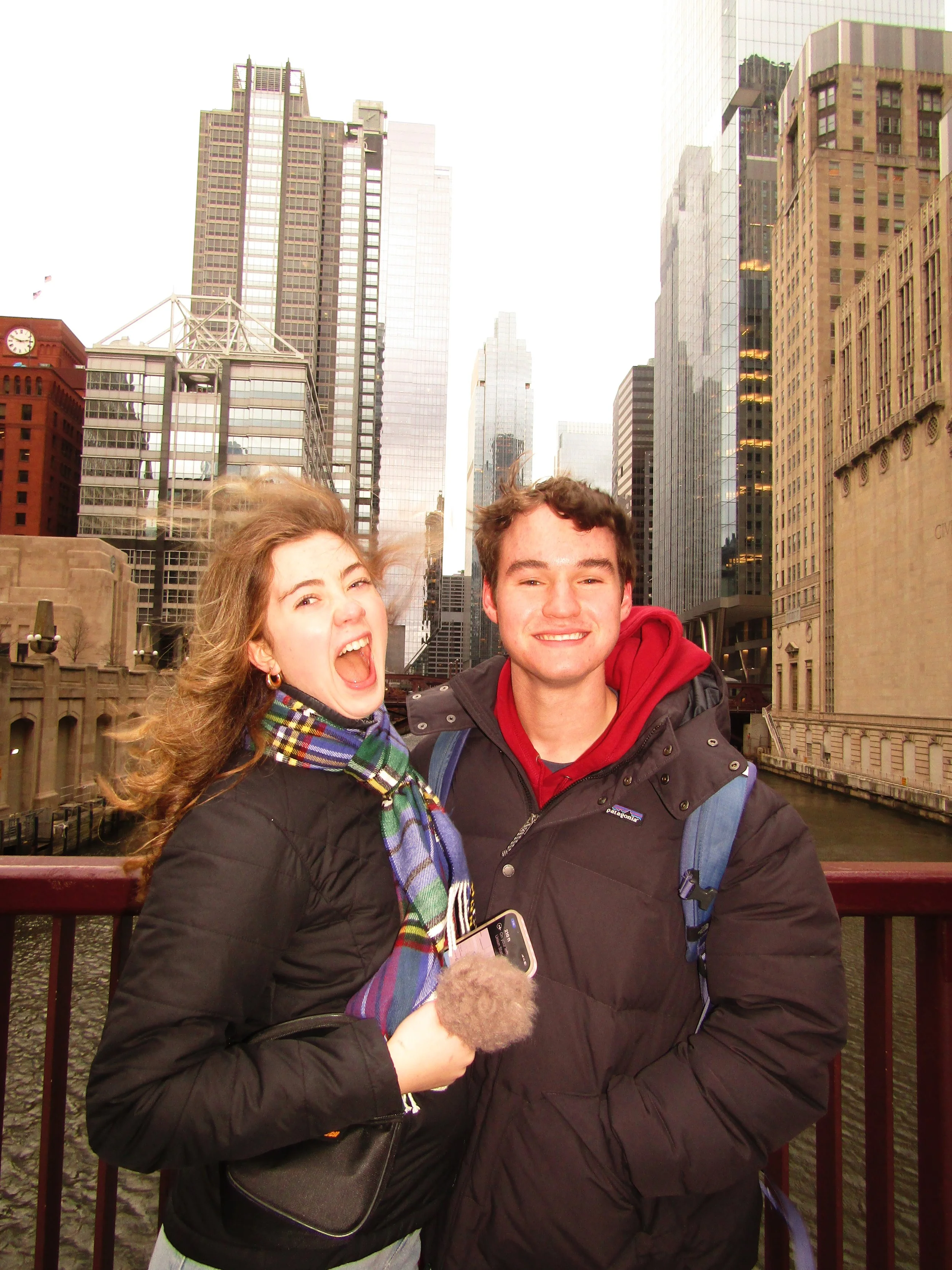 Two young adults, a woman and a man, standing on a bridge in front of a city skyline with tall skyscrapers, water, and a cloudy sky.