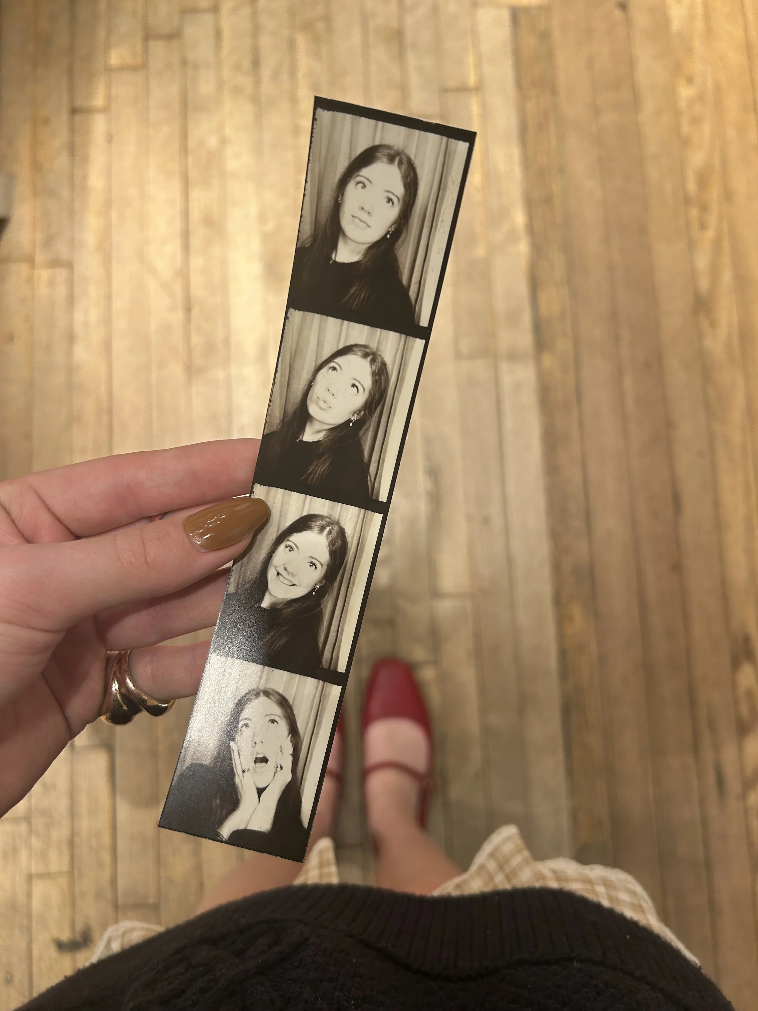 A hand holding a four-photo strip of black-and-white photo booth pictures of a woman making different facial expressions, with a wooden floor background.