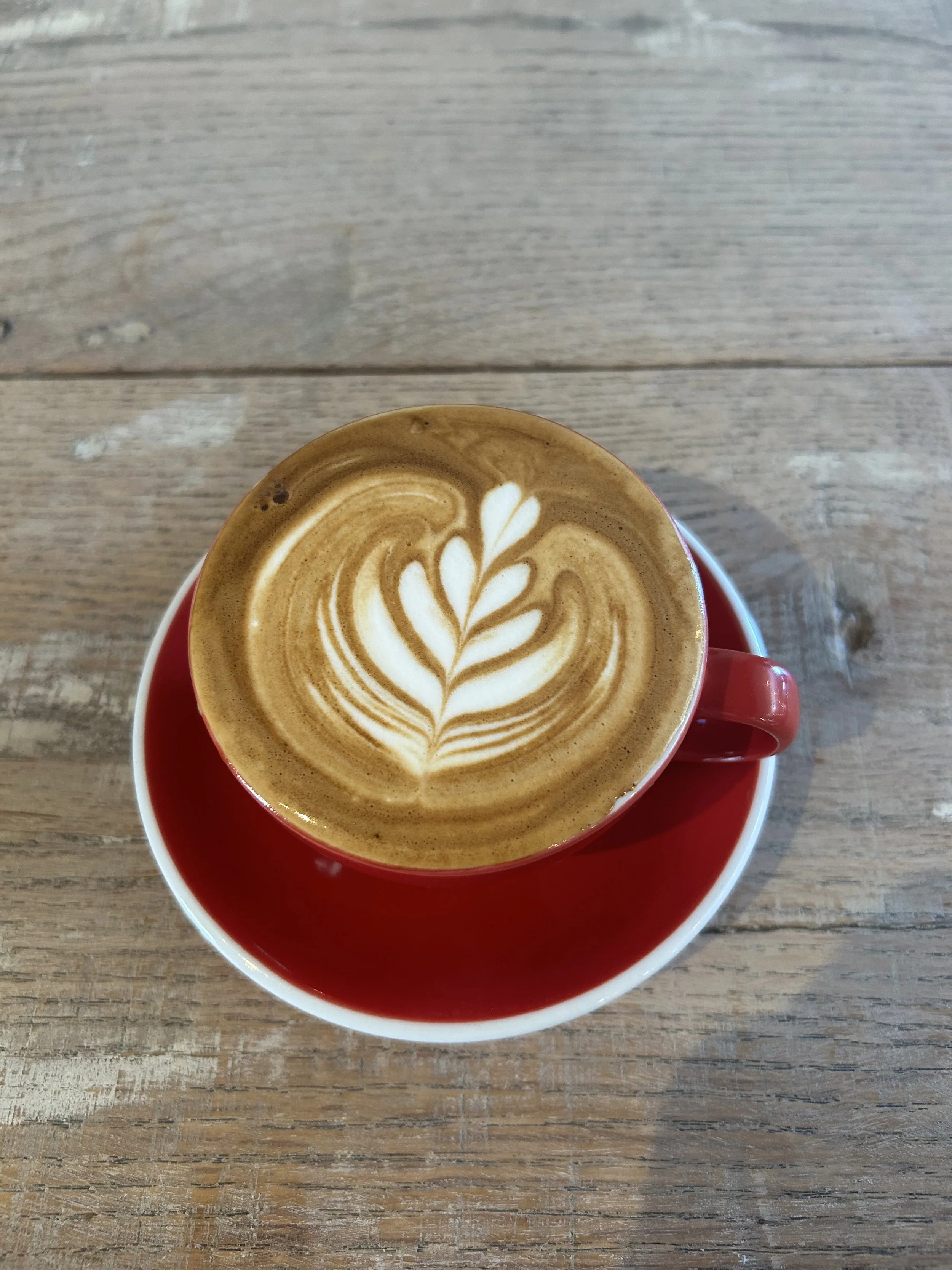 A latte in a red cup with a leaf-shaped foam art design on top, sitting on a matching red saucer on a wooden table.
