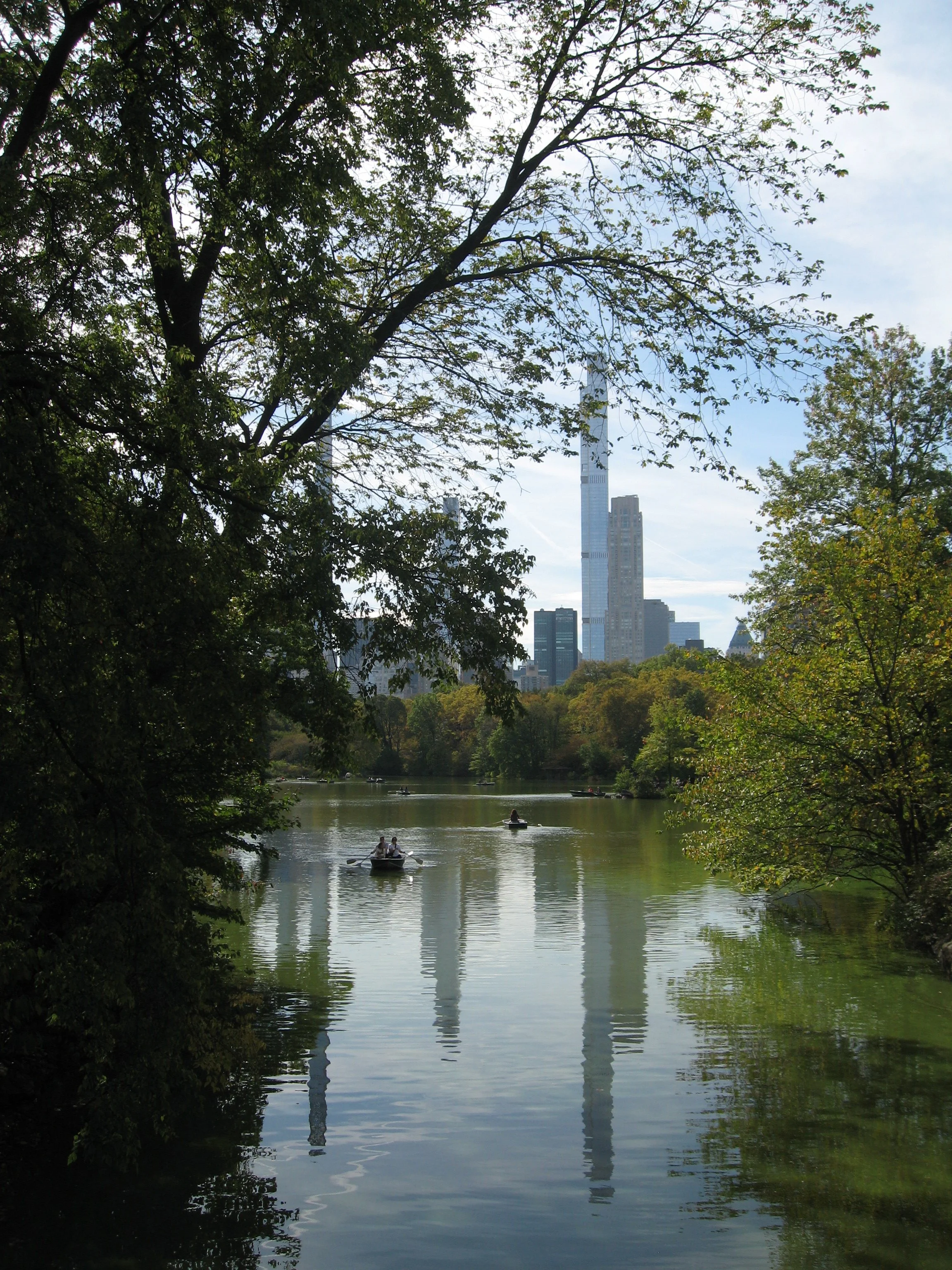 A peaceful scene of boats on a river in a park with lush trees, with a city skyline featuring tall skyscrapers in the background.