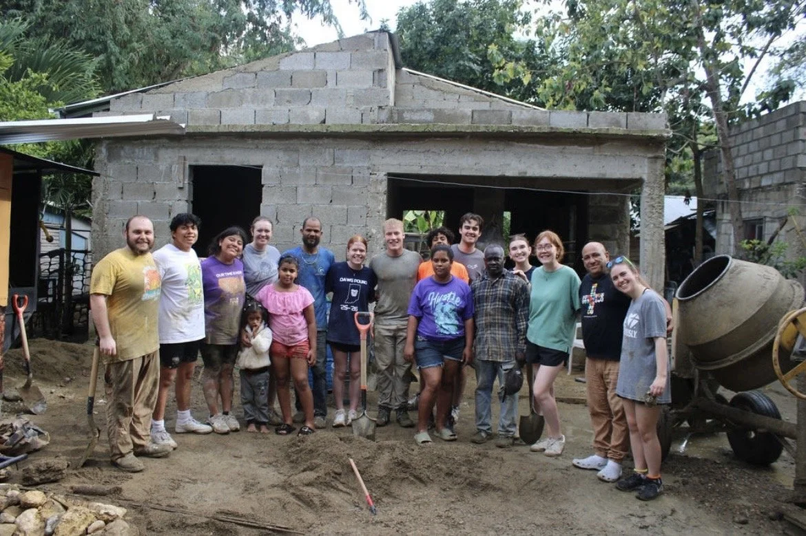 Group of people standing in front of a construction site with an unfinished brick house, some holding shovels, on a dirt ground surrounded by trees.
