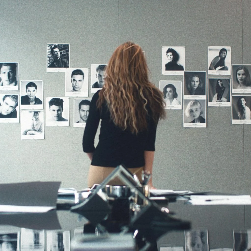 A woman with long, wavy hair wearing a black top looks at black-and-white headshots of various people displayed on a gray wall in an office or studio setting.