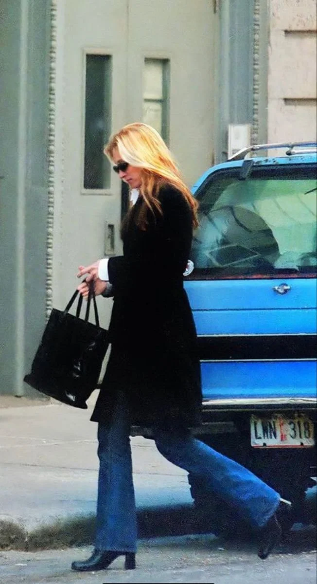 A woman with blonde hair wearing black sunglasses, a black coat, and blue jeans, walking on the street carrying a black bag next to a blue vehicle.
