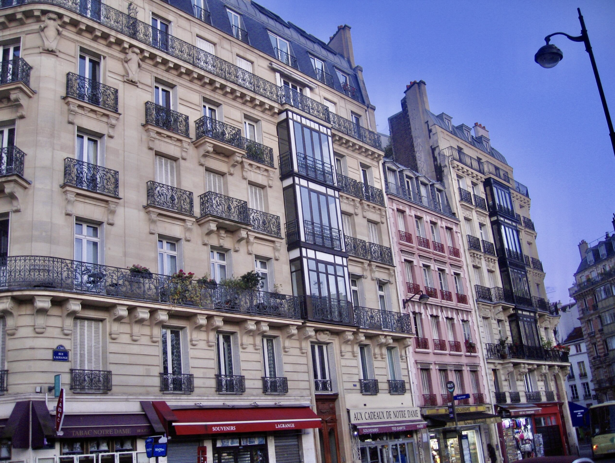 Parisian street scene with multi-story buildings featuring classic wrought-iron balconies and modern glass extensions, storefronts at street level, and a street lamp against a blue sky.