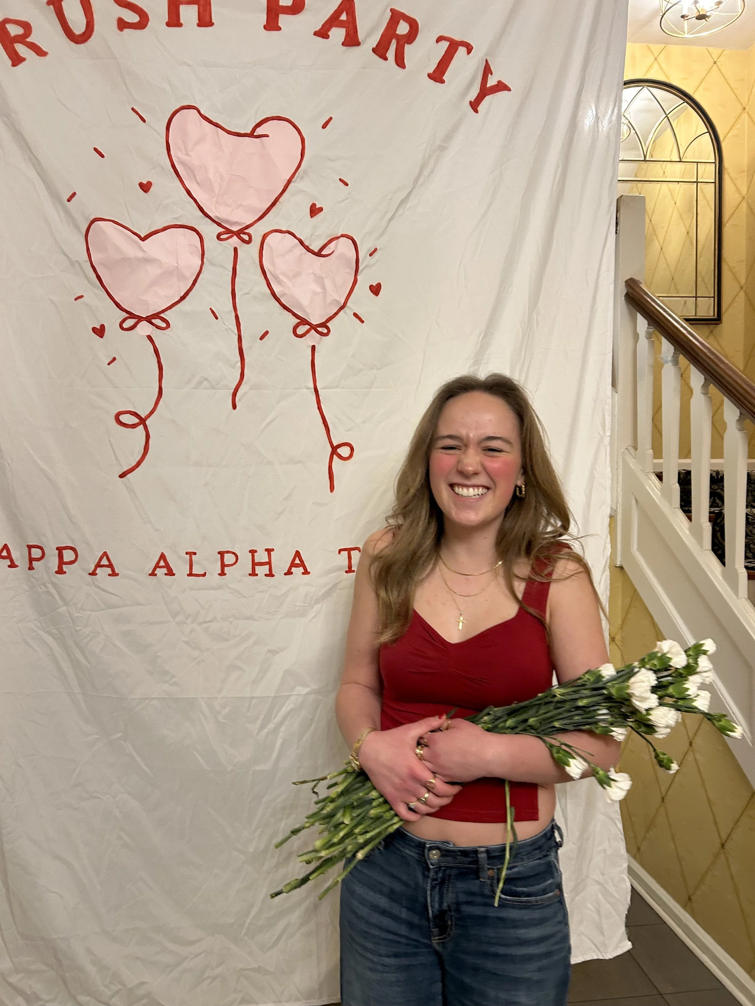 A young woman with long blonde hair, wearing a red top and jeans, smiling and holding a large bouquet of white flowers, standing in front of a white backdrop with red balloons and the words 'Rush Party' and 'Alpha' in red letters.