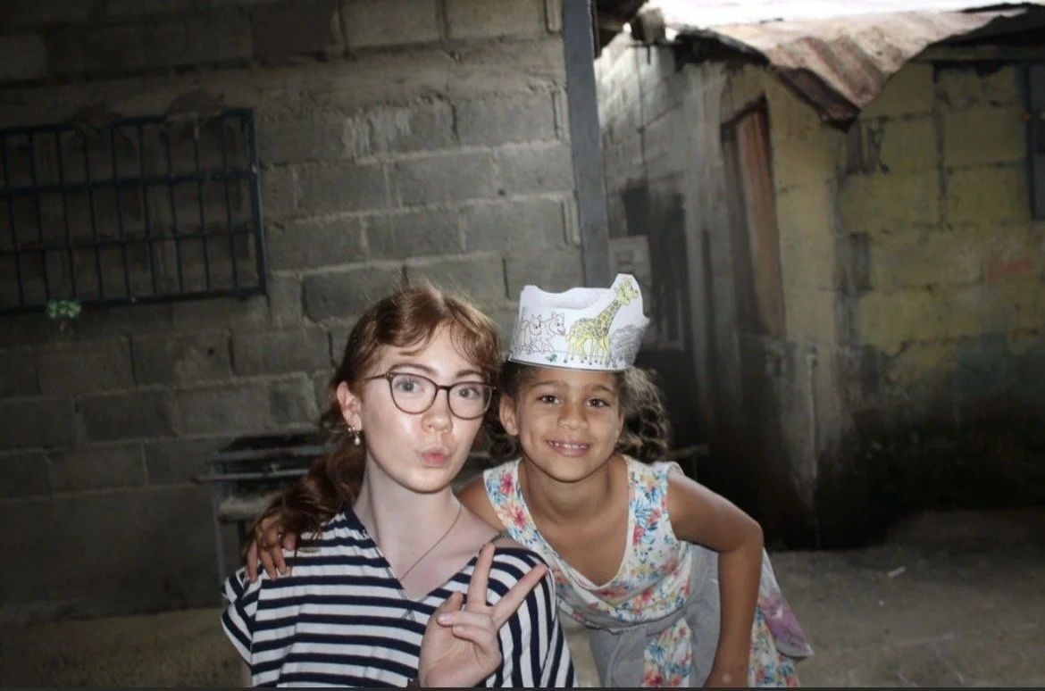 Two young girls smiling and posing for the camera outdoors. One girl is wearing glasses and making a peace sign, while the other girl is wearing a paper crown with animal drawings.