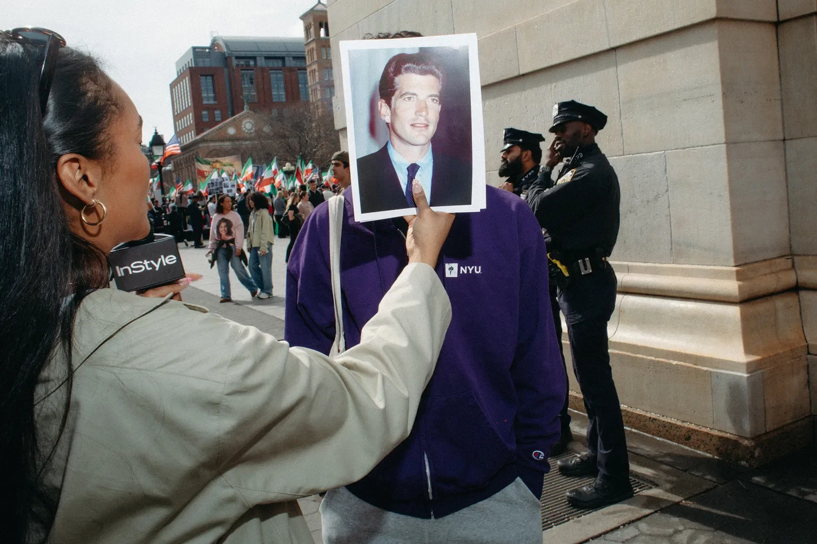 A woman holding a photo of a man in front of a man wearing a purple jacket, with police officers in the background during a protest or rally.