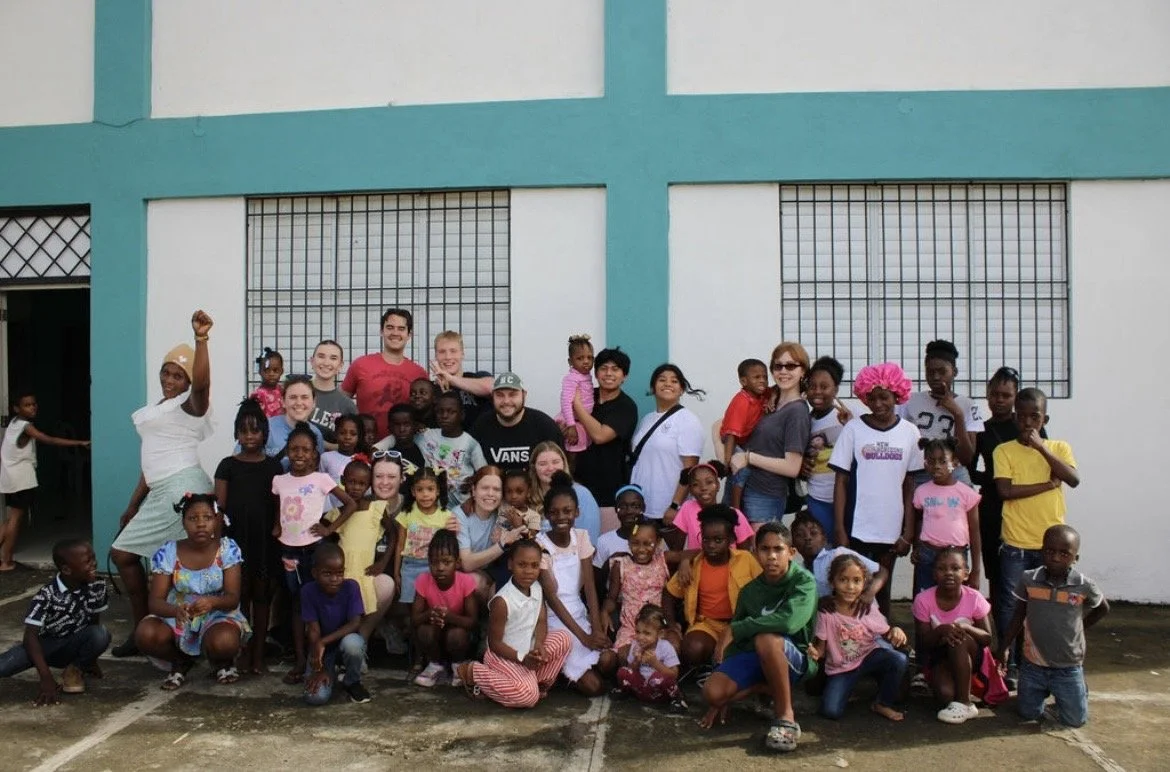 Group of children and adults posing outside a building with blue beams and barred windows.