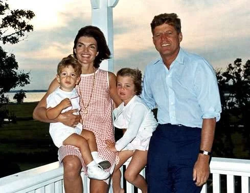 A family of four, including a woman, a man, and two children, standing outdoors on a white porch with a wooden railing near a body of water, during sunset.