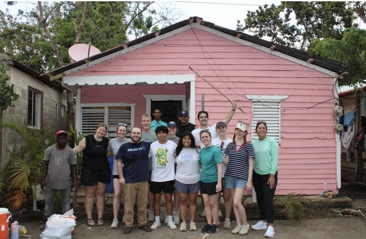 Group of people standing in front of a pink wooden house, smiling and posing for a photo.