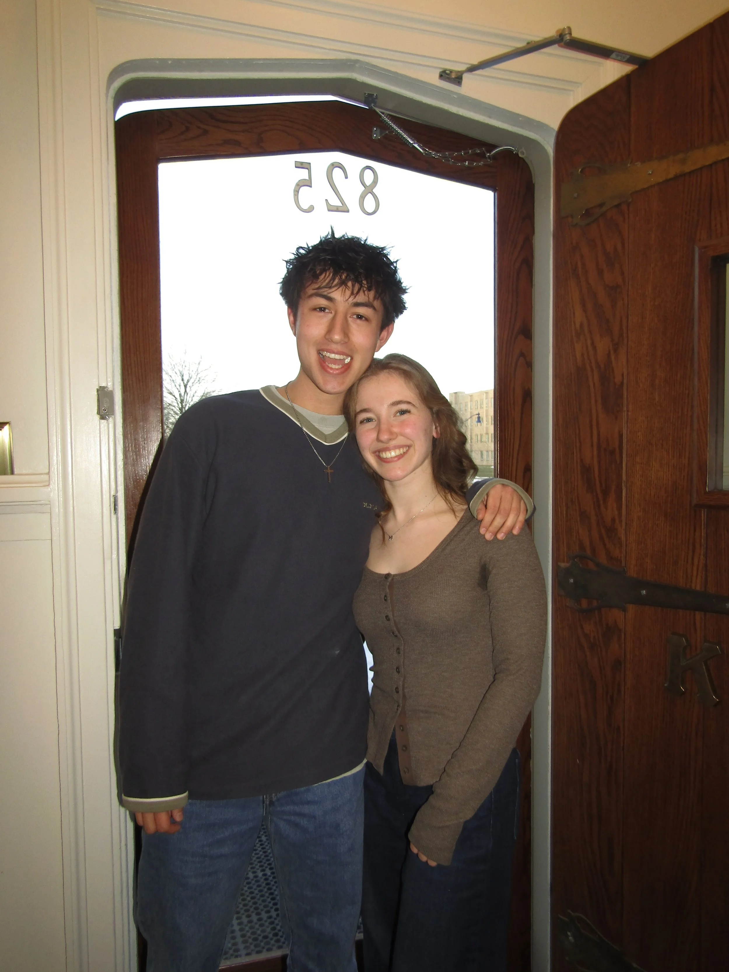 A young man and woman standing together indoors, smiling, with the young man having his arm around the woman's shoulder near a wooden door.