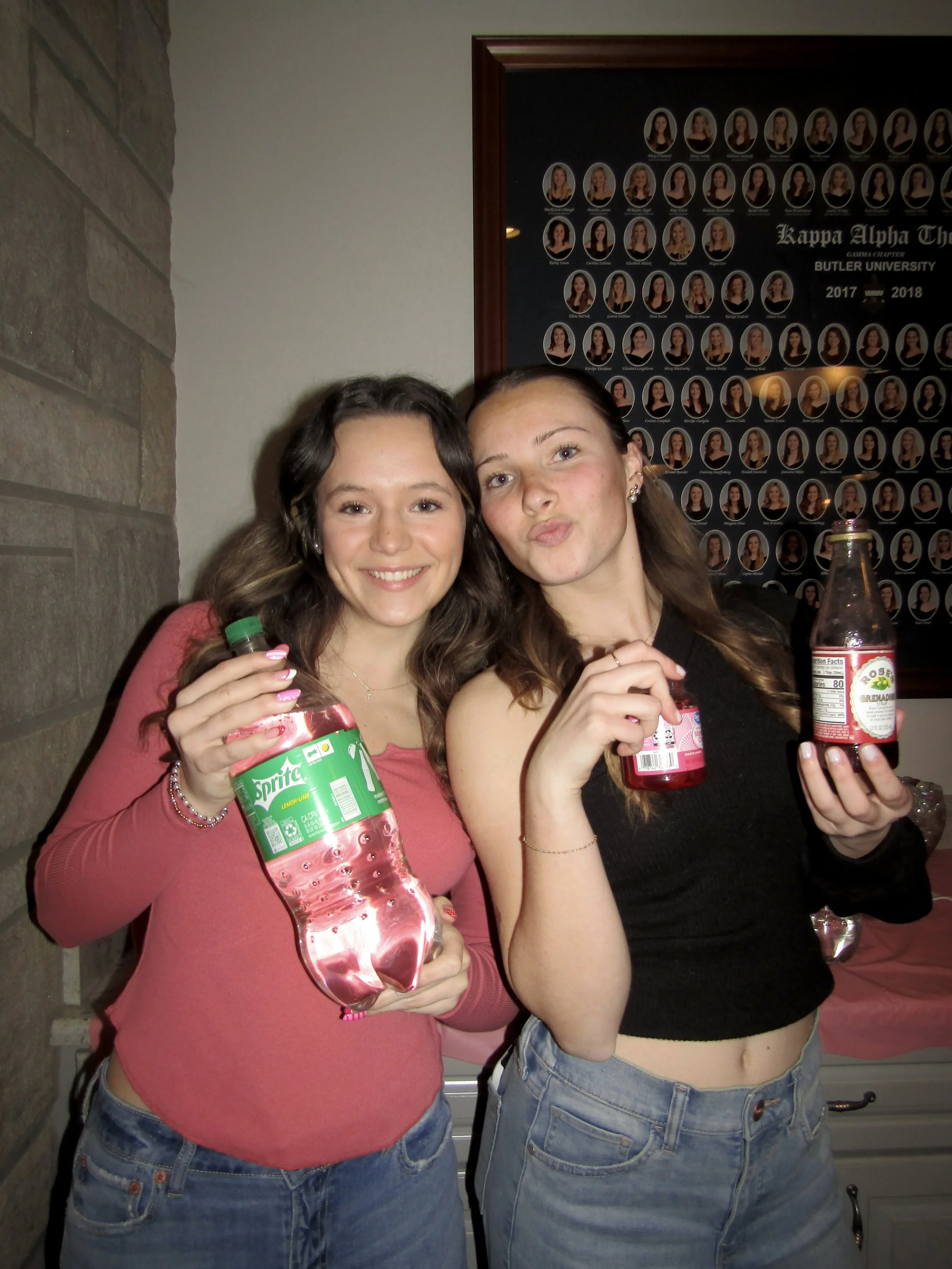 Two young women smiling and holding bottles of soda while standing indoors. One is holding a Sprite bottle, and the other is holding a Coca-Cola bottle. They are in front of a commemorative display with graduation photos.