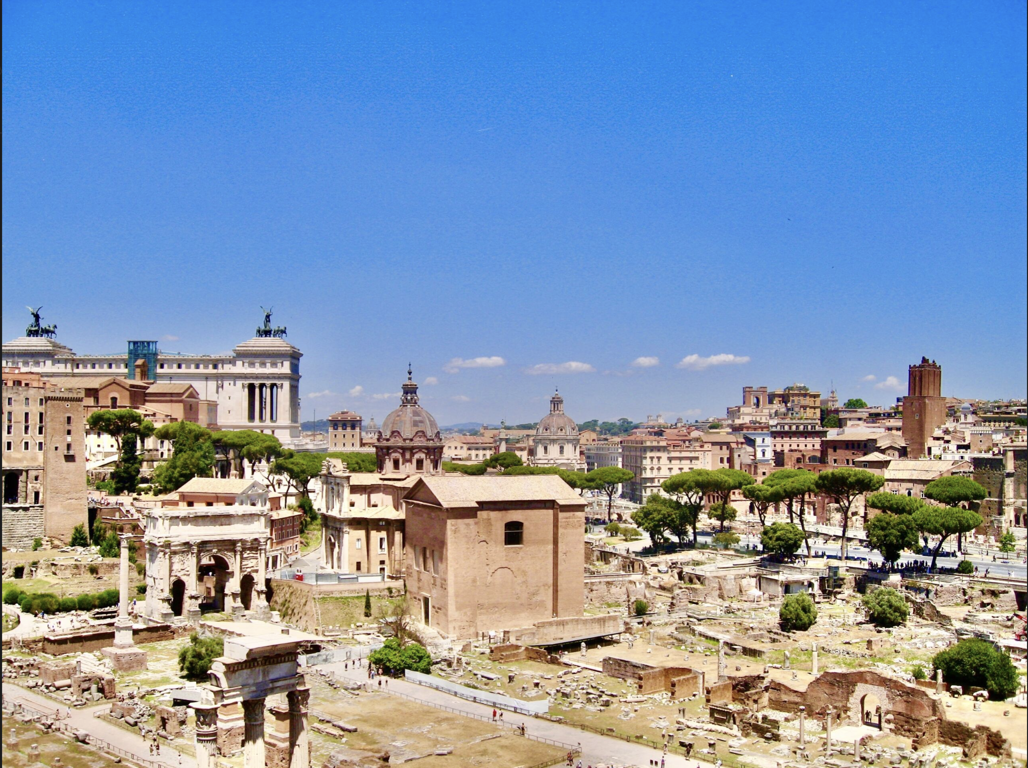 A panoramic view of ancient ruins and historic buildings in Rome, Italy, under a clear blue sky with some clouds.