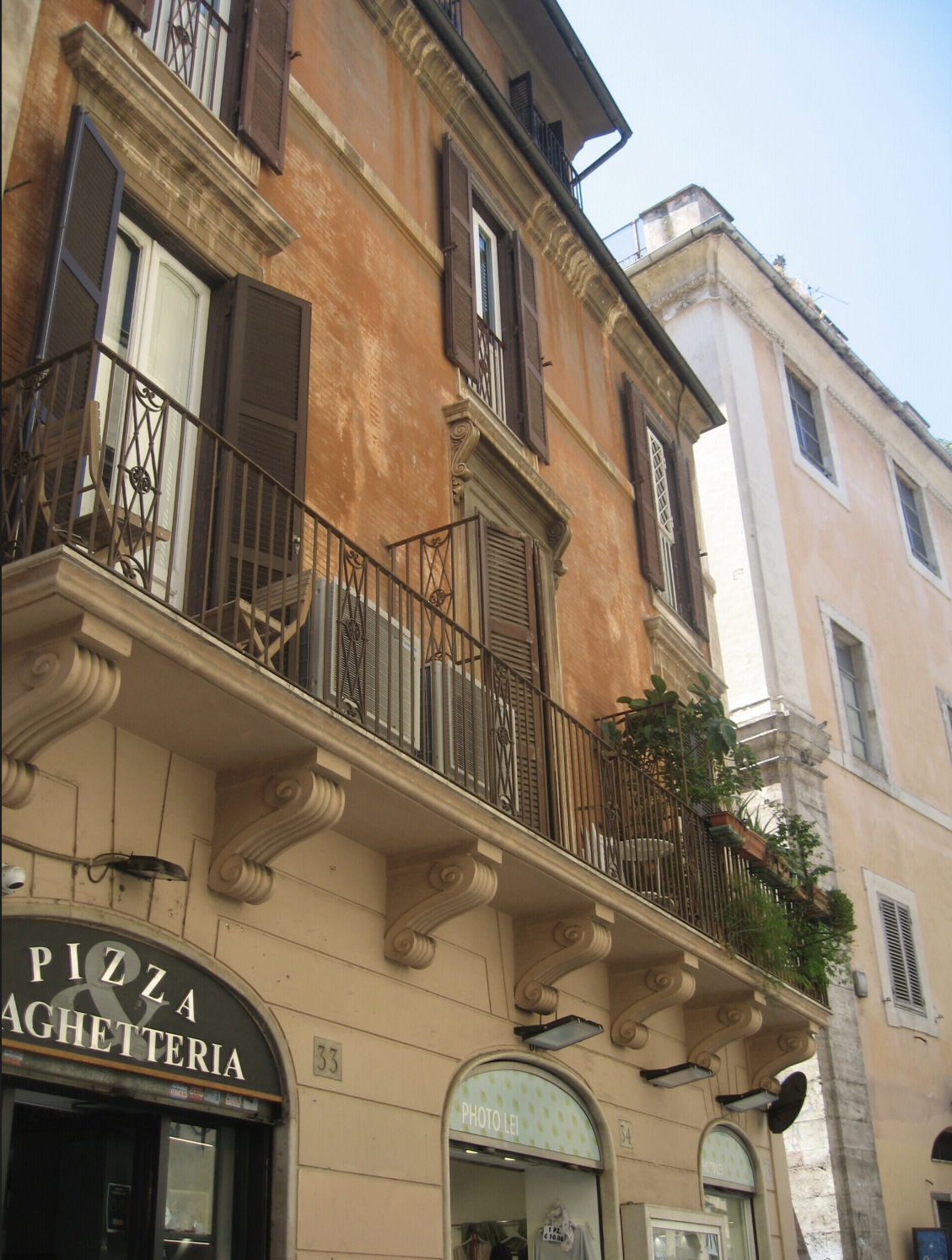 Buildings with orange and beige facades, small windows with shutters, balconies with plants, and a storefront with Italian words 'Pizza' and 'Pasticceria'.