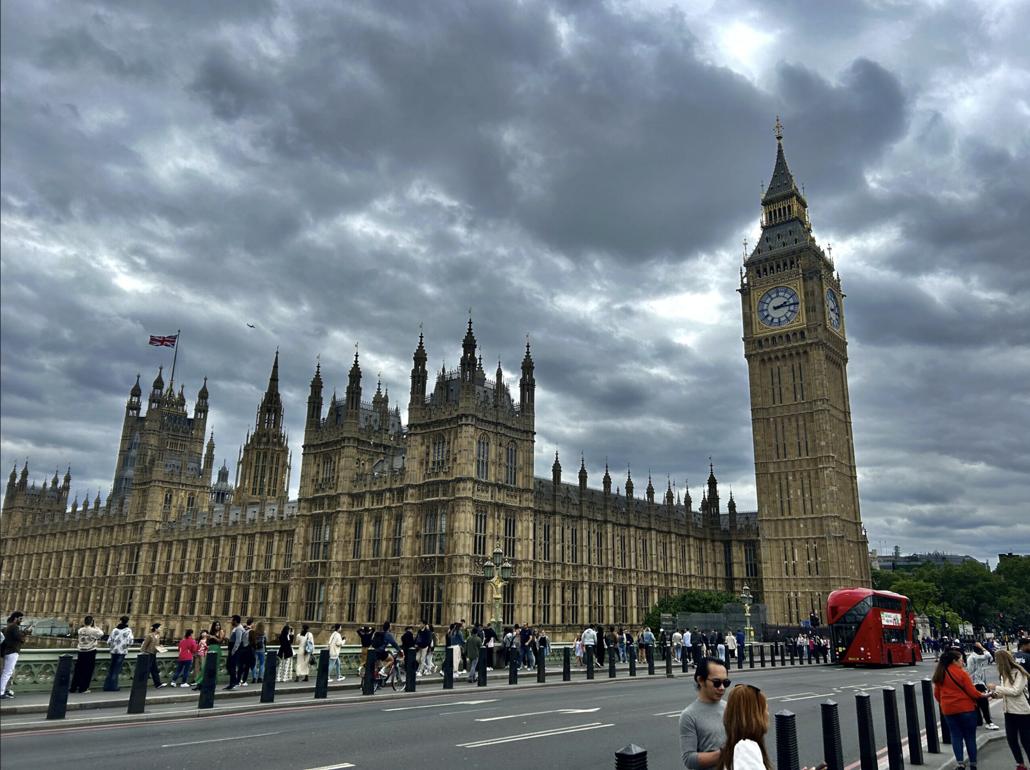 View of the Palace of Westminster in London, featuring the Big Ben clock tower, with cloudy skies overhead and people walking along the nearby street.