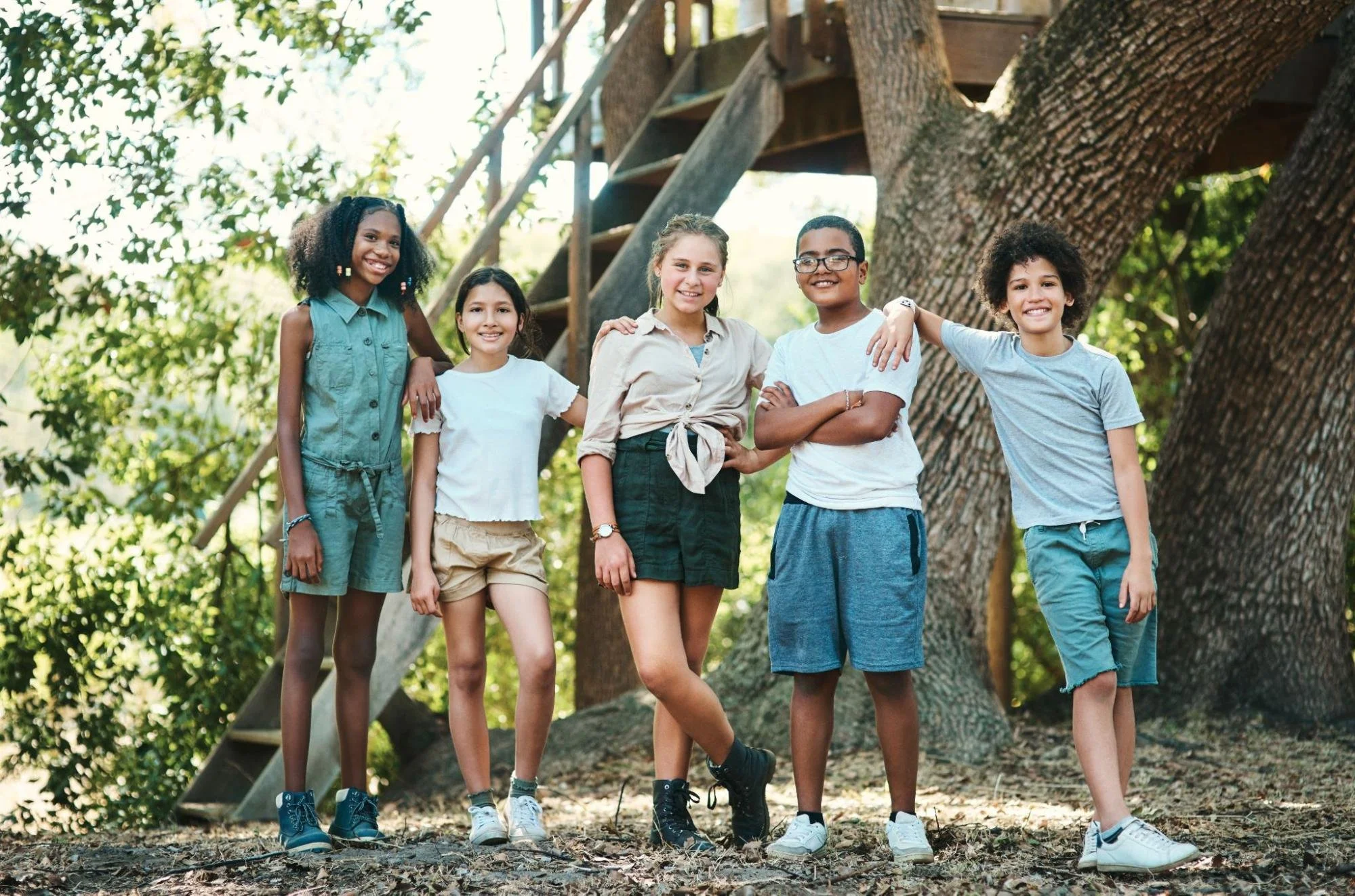 A group of teenagers standing next to a tree at summer camp happily smile at the camera. 