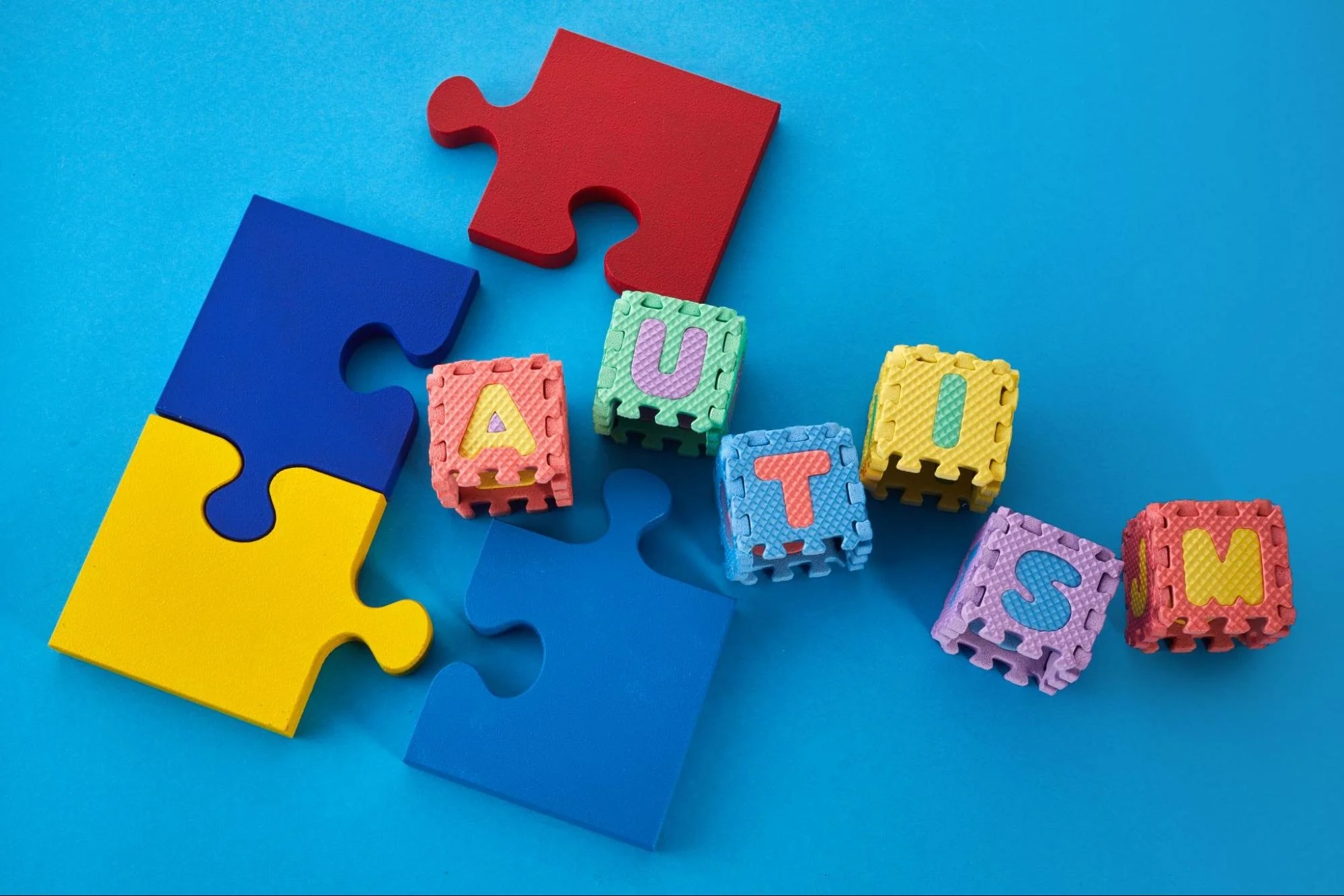 Colorful puzzle pieces around foam blocks that spell "Autism" arranged on a bright blue background.