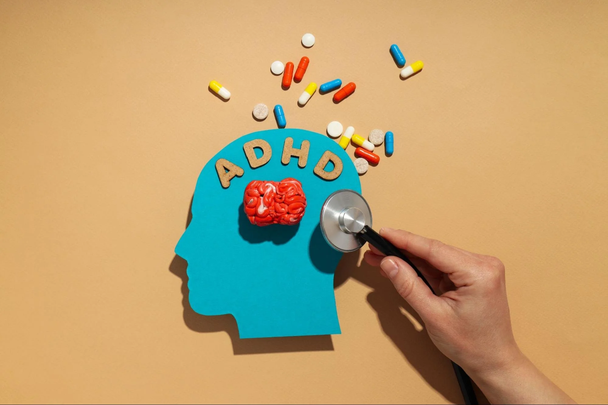 A cutout of a human head with "ADHD" written on it is shown with a small brain model, scattered pills, and a stethoscope.