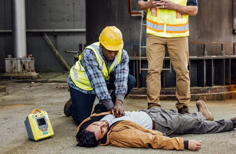 A person in a yellow safety helmet and vest performs CPR on an unconscious person lying on the ground, with an AED device nearby.
