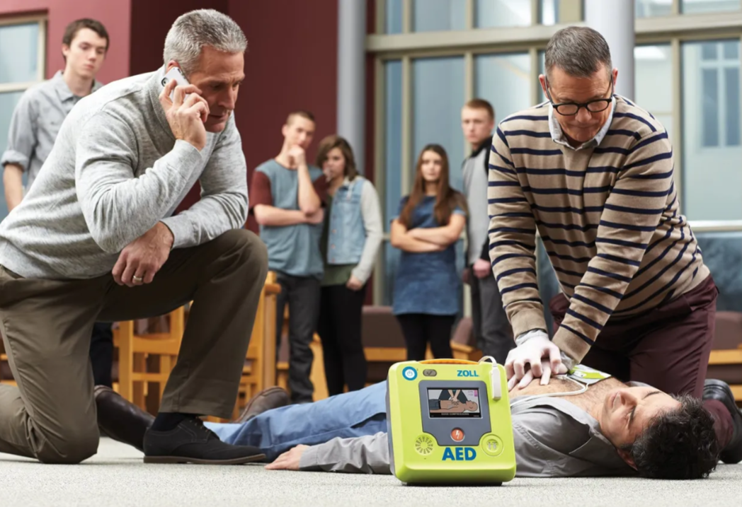 Two men perform CPR on a man lying on the floor during a training session, with a defibrillator nearby, while several people watch.