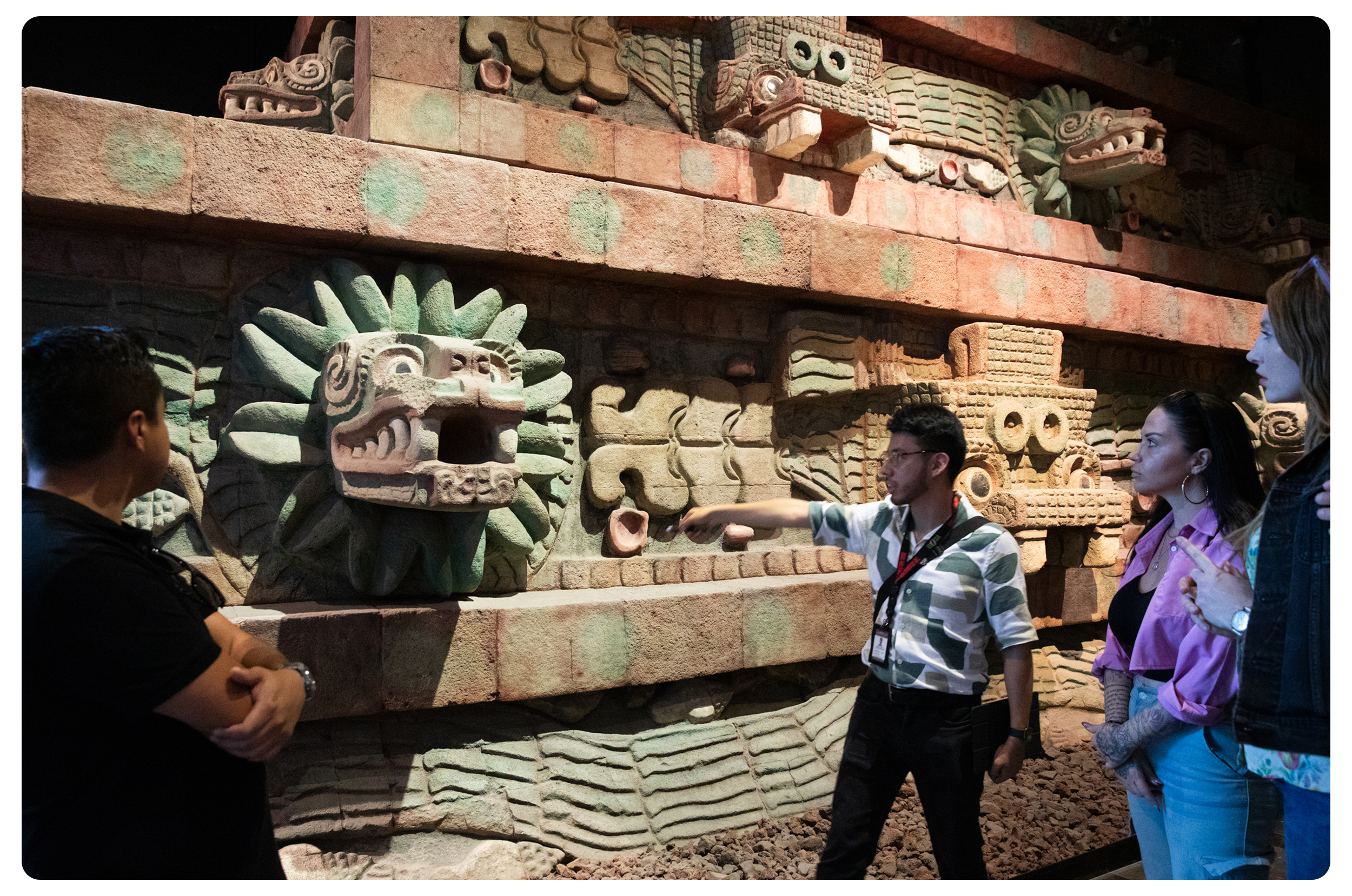 A guide explains ancient stone carvings, including a jaguar head with a large mouth and detailed features, to three women inside a museum.