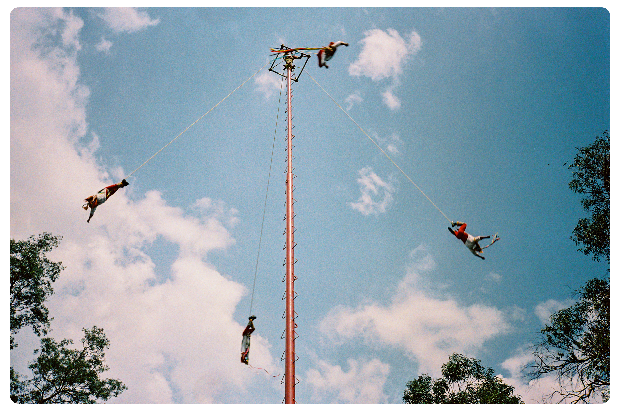 People participating in a traditional Mexican game called 'Tlachtli' involving balancing on a tall pole with swinging ropes and hanging figures.