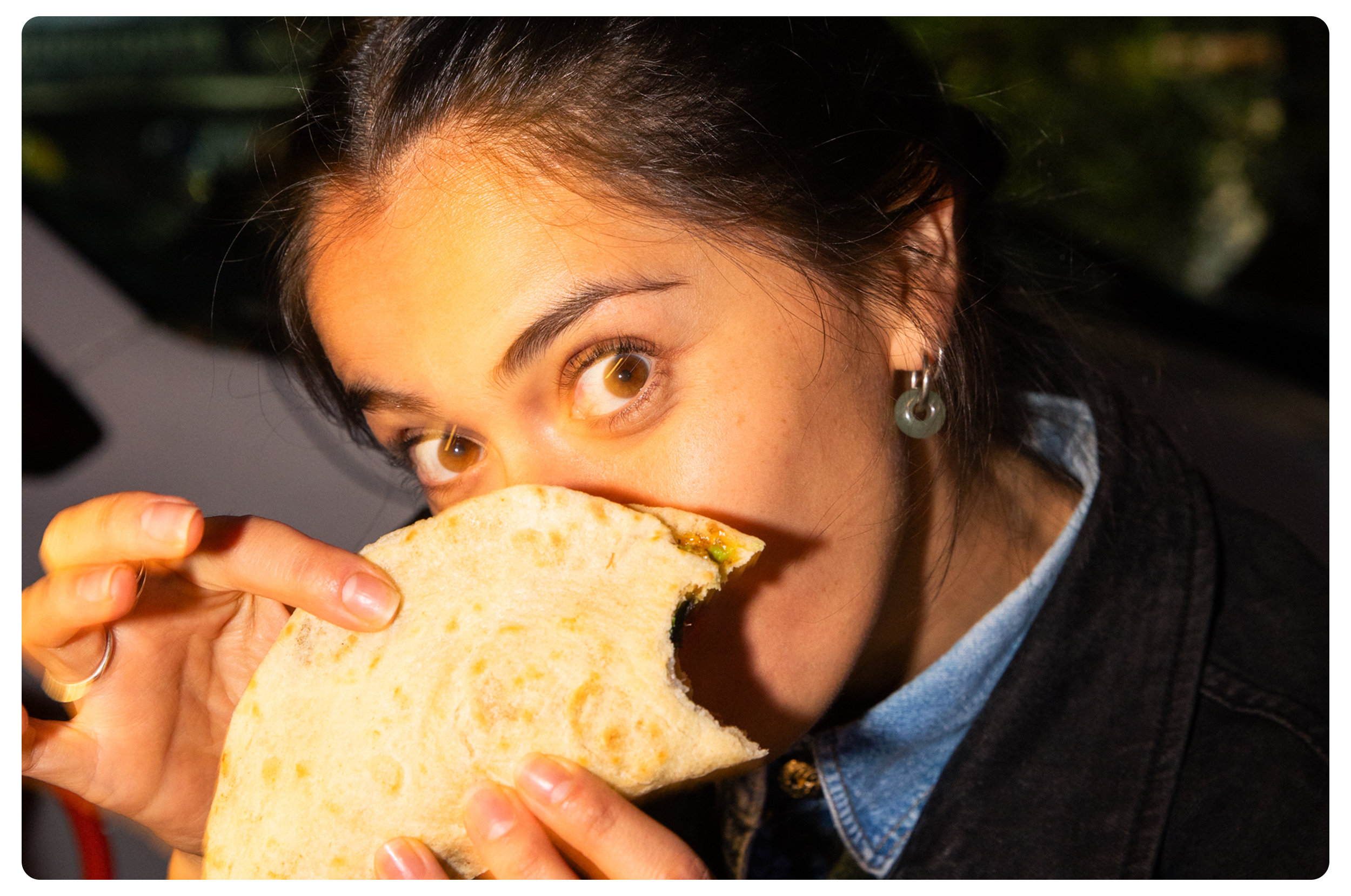 A woman with brown hair and hoop earrings is eating a partially bitten sandwich or pita with filling inside.