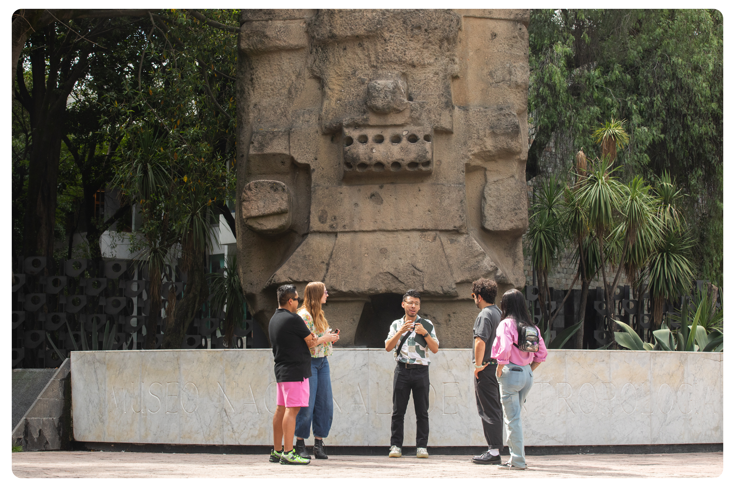 Group of five people standing in a circle and talking in front of a large stone sculpture at Museo Nacional de Antropología, Mexico City, surrounded by trees and plants.