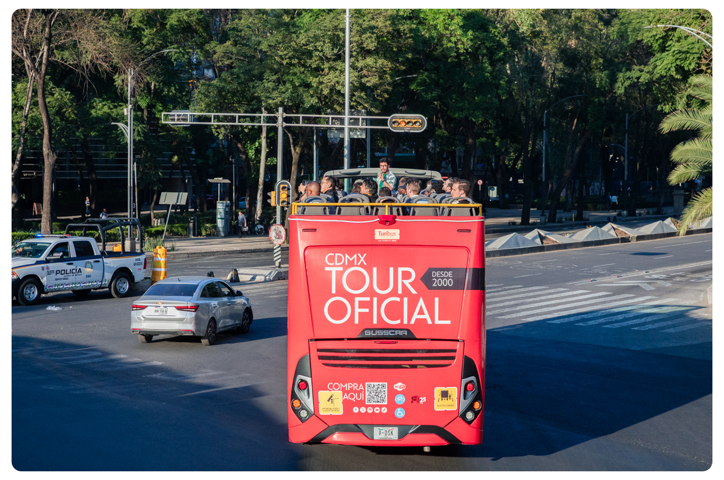 Open-top red tour bus with passengers on a city street in Mexico City, with police car and cars around, and green trees in the background.