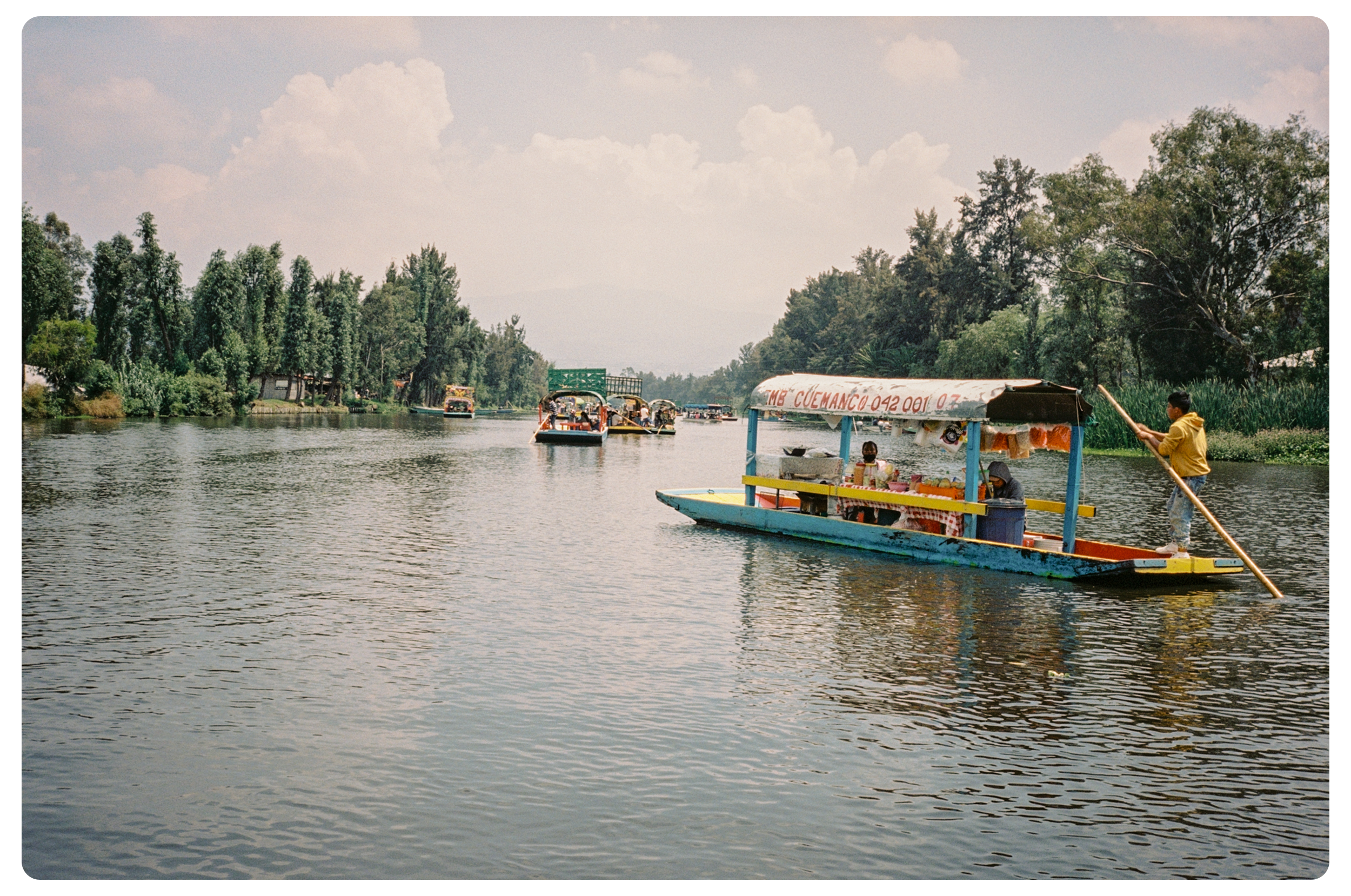 Colorful boat with a canopy on a river, carrying people and goods, with trees on the riverbanks and other boats in the background under a partly cloudy sky.