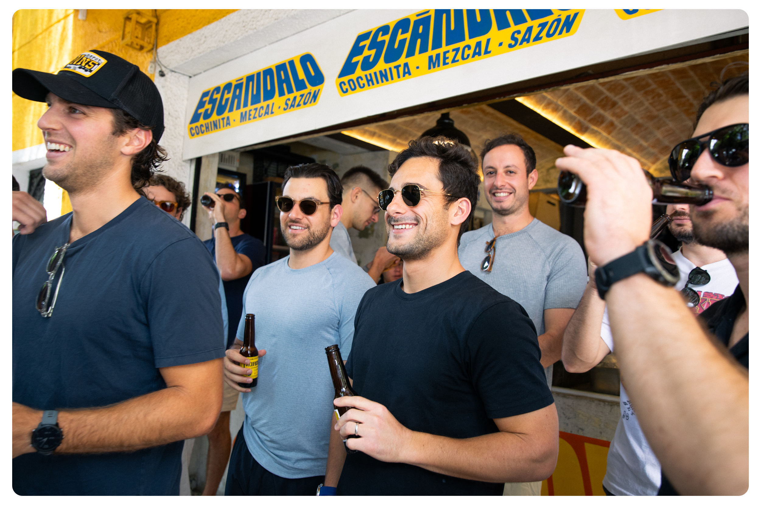 Group of men at a social gathering, smiling and holding drinks, outside a bar with a sign that reads 'Escándalo' in Spanish, advertising cochinita, mezcal, and a season.
