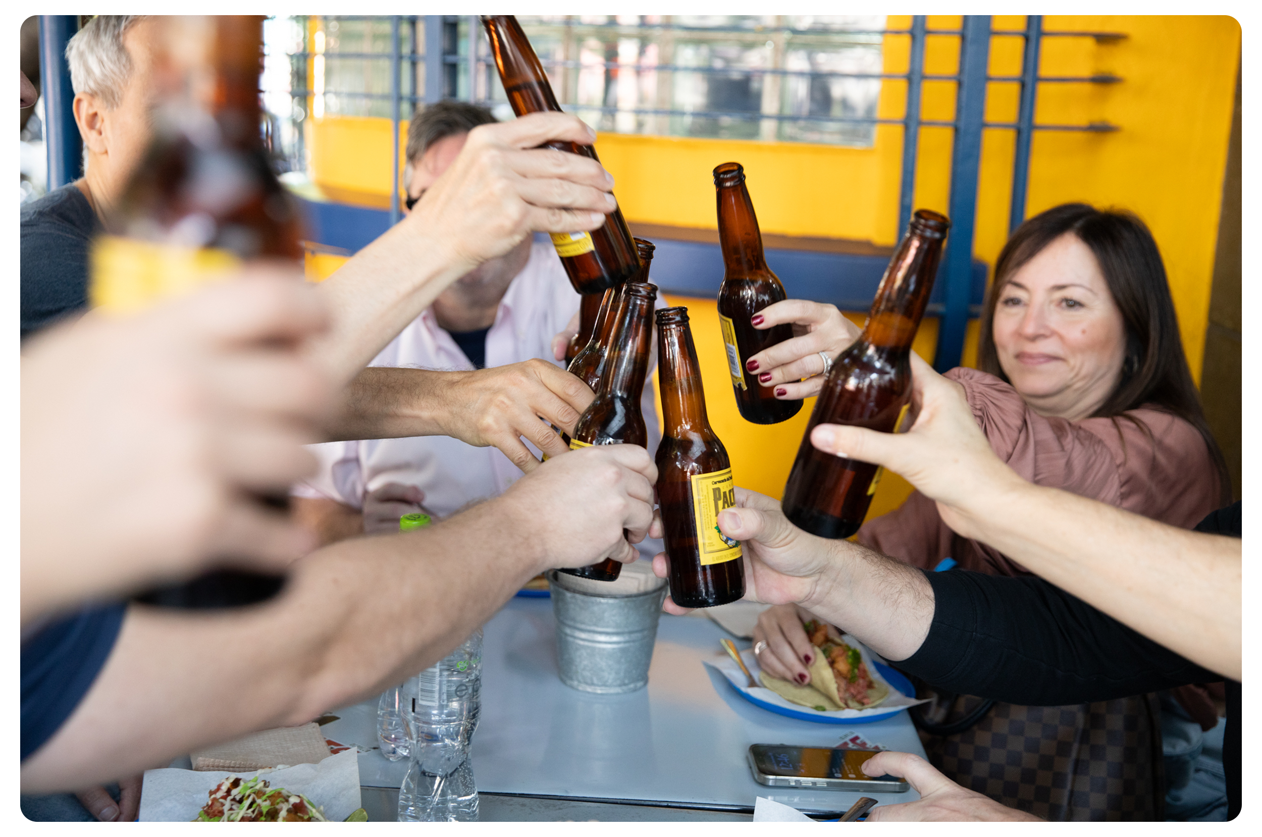 A group of people raising beer bottles in a toast at a table on a patio with a yellow and blue wall in the background, with some food and drinks on the table.