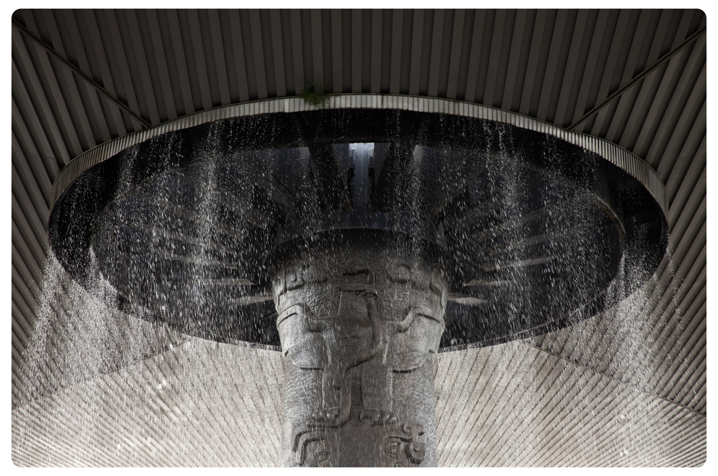 Close-up of an architectural feature with water cascading from a circular structure, beneath a large, curved roof with vertical paneling.